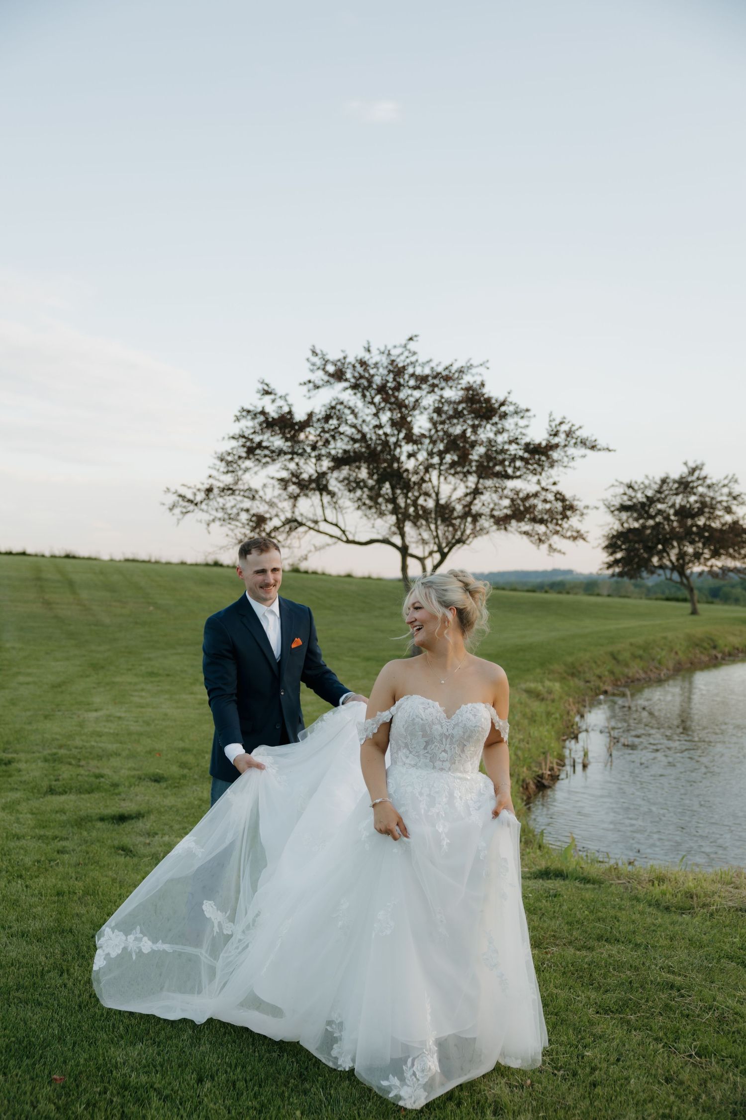 Bride in flowing off-shoulder lace gown walks with groom in navy suit along scenic countryside pond.