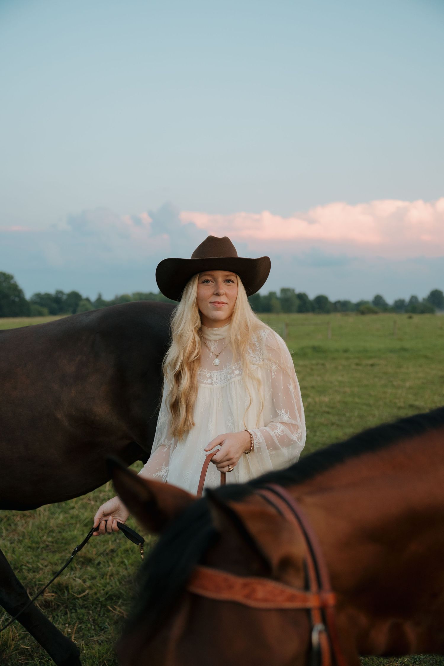 Blonde woman in cowboy hat and white dress standing with brown horses in a green field at sunset.