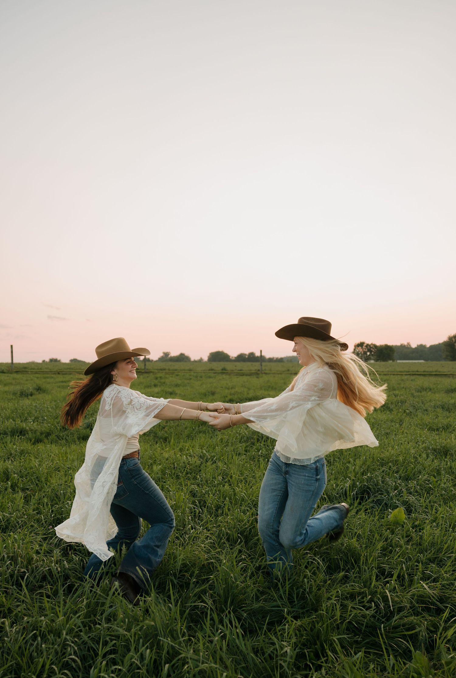 Two women in cowboy hats and white blouses hold hands and spin joyfully in a green field at sunset.