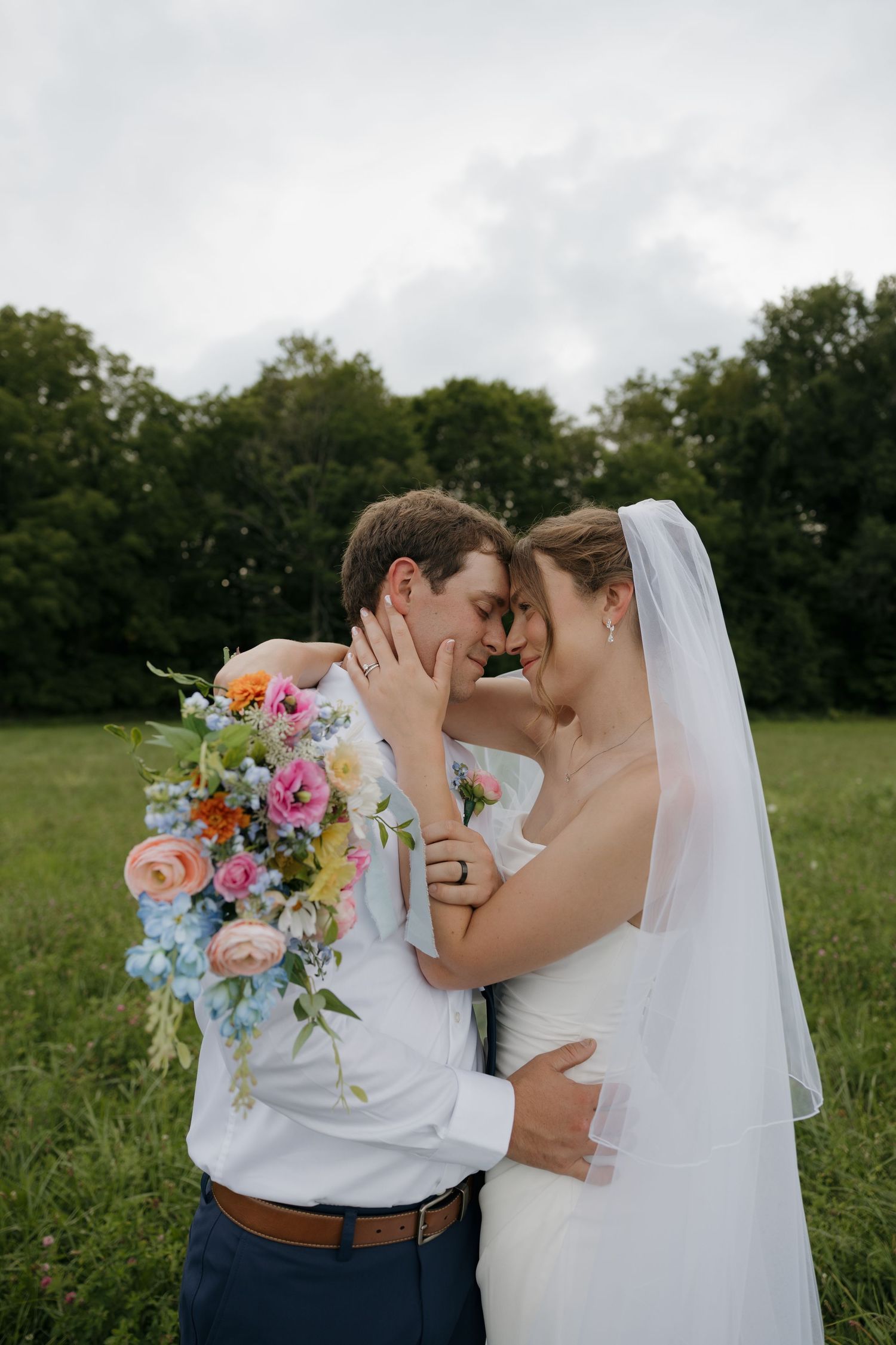 Bride and groom share a romantic forehead touch outdoors, bride holding colorful floral bouquet in a green field.