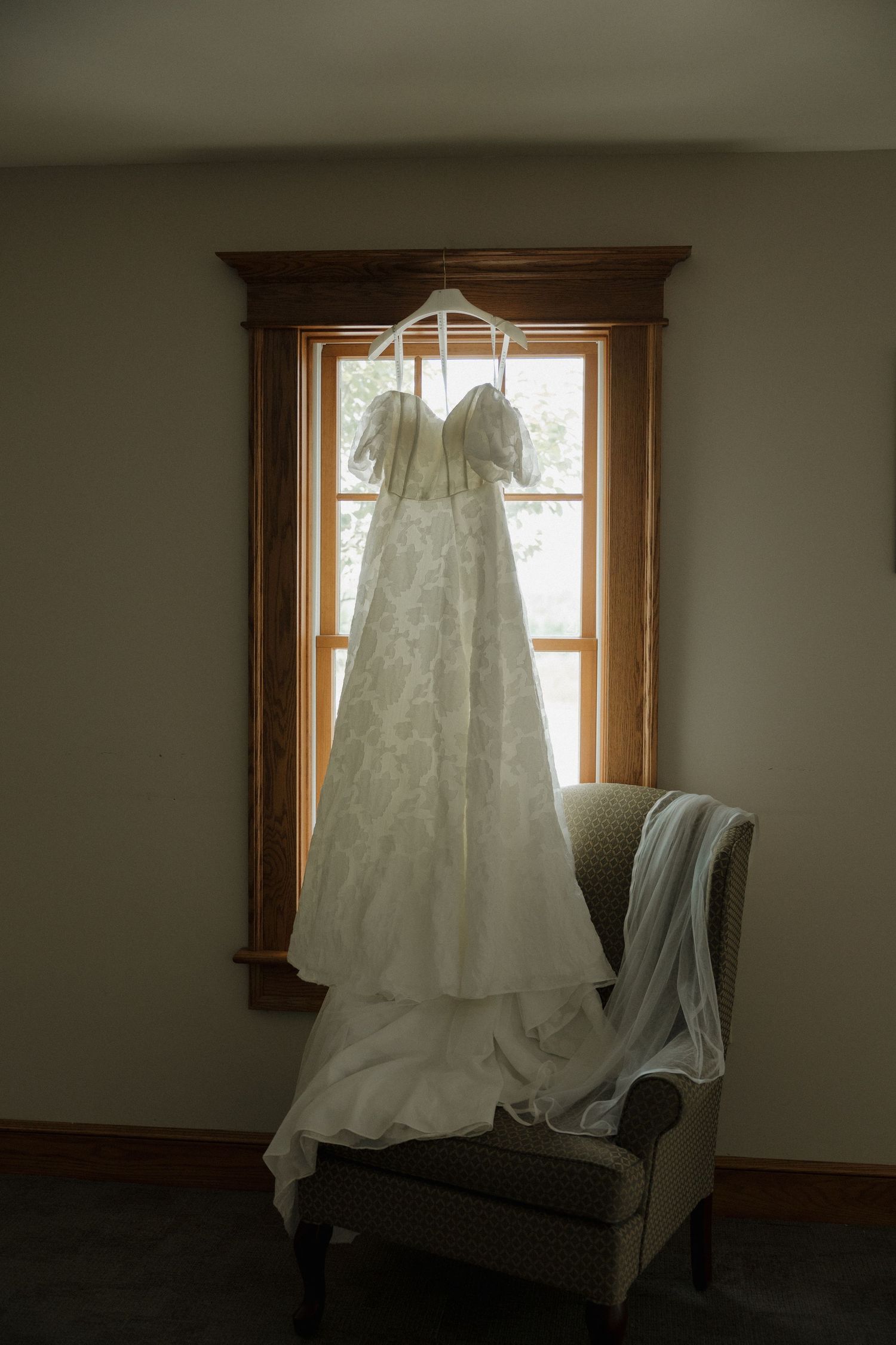 White off-shoulder lace wedding gown hanging on a hanger in front of a wooden-framed window.