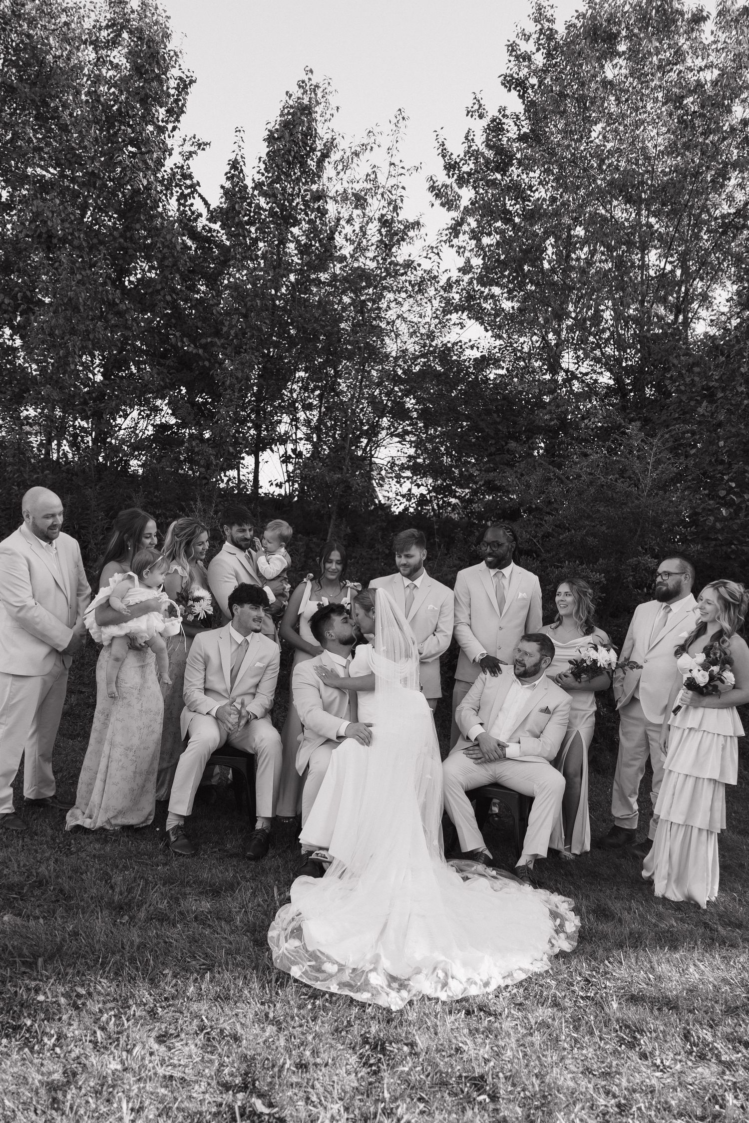 Black and white wedding photo of bride and groom surrounded by wedding party on a grassy lawn with trees in background.
