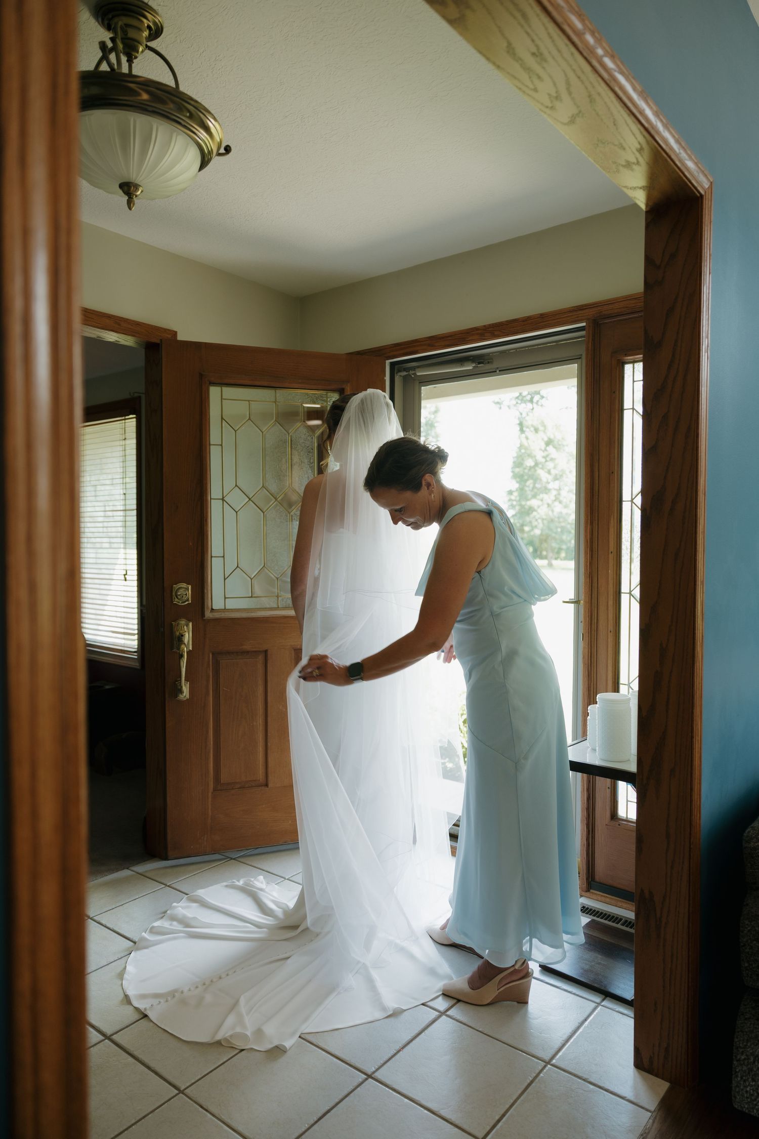 Bridesmaid in blue dress adjusting the train of a bride's white wedding gown in a sunlit home entryway.