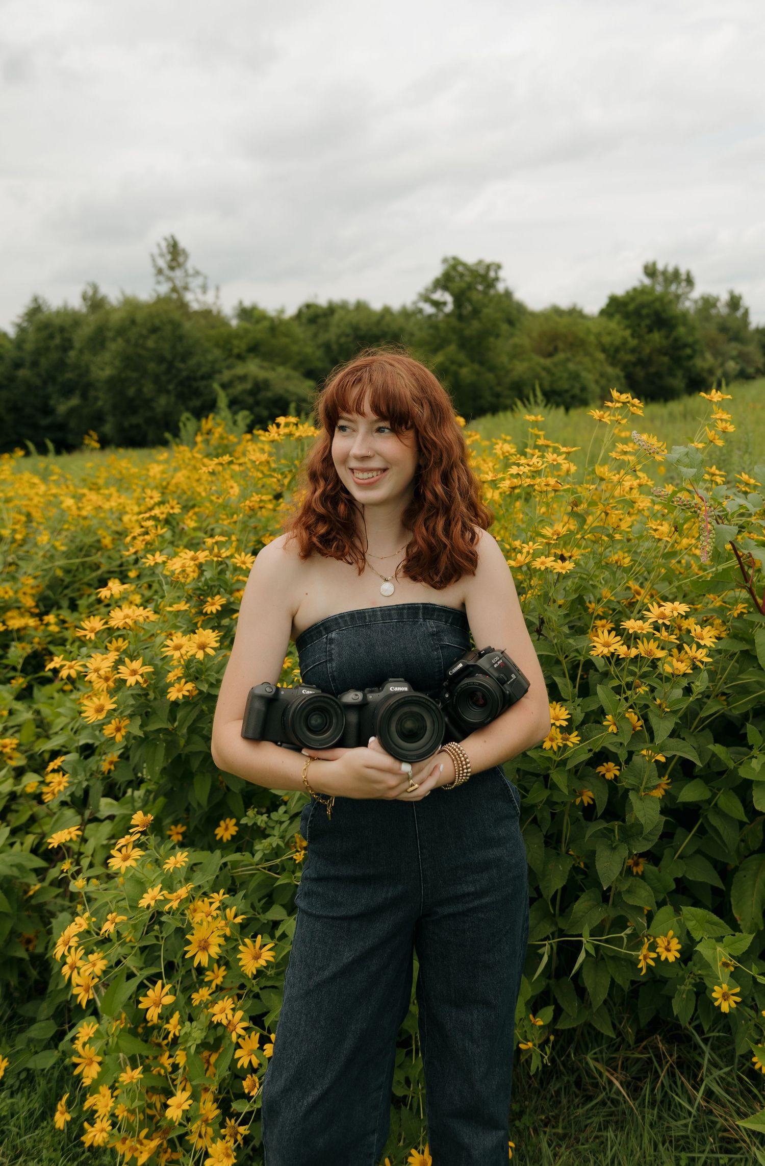 A young woman with red hair holds cameras while standing in a field of yellow wildflowers on a cloudy day.
