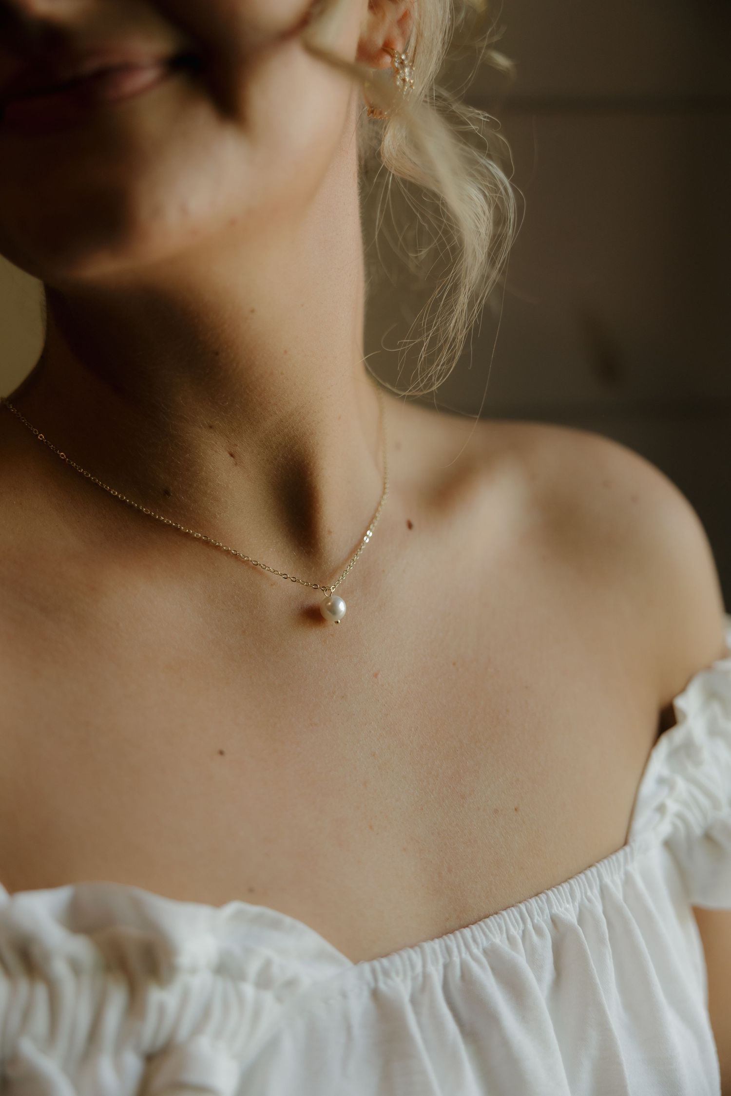 Close-up of a bride wearing a delicate pearl pendant necklace on a gold chain with an off-shoulder white dress.