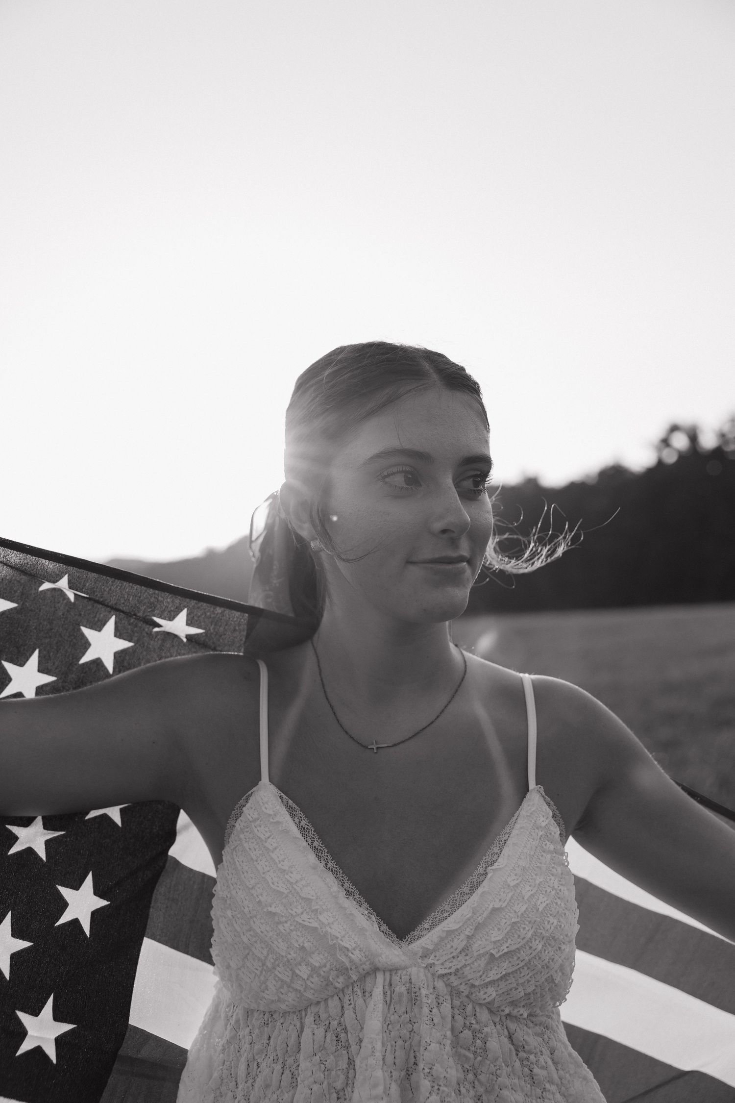 Black and white photo of a young woman holding an American flag behind her shoulders outdoors at sunset.