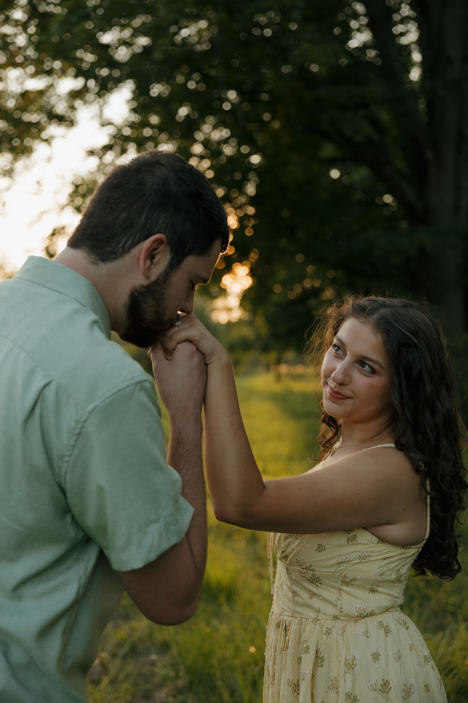 A couple shares a romantic moment at golden hour, the woman in a floral dress smiling as her partner kisses her hand.
