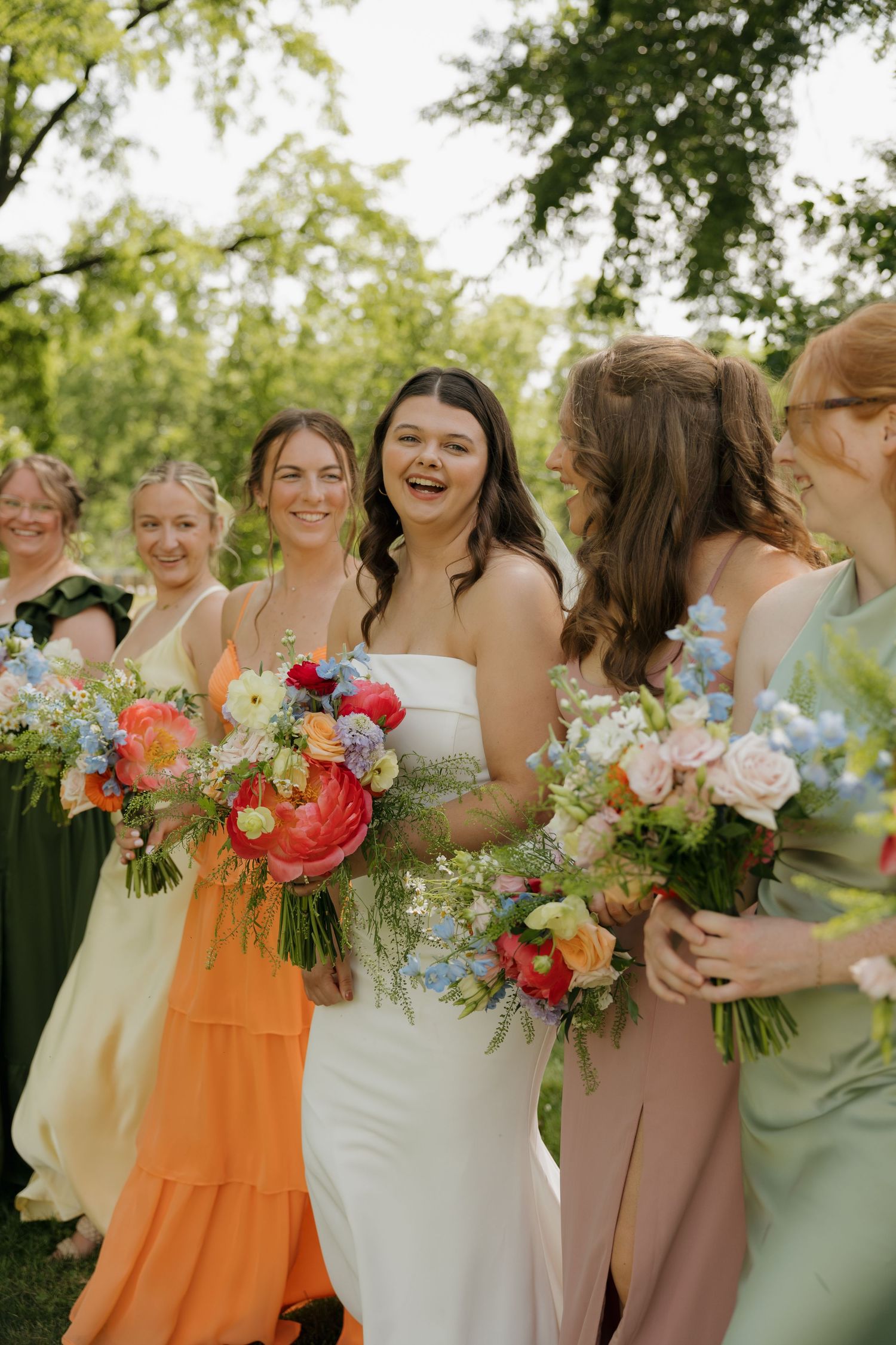Bride in white gown laughs with bridesmaids in colorful mismatched dresses holding vibrant floral bouquets outdoors.