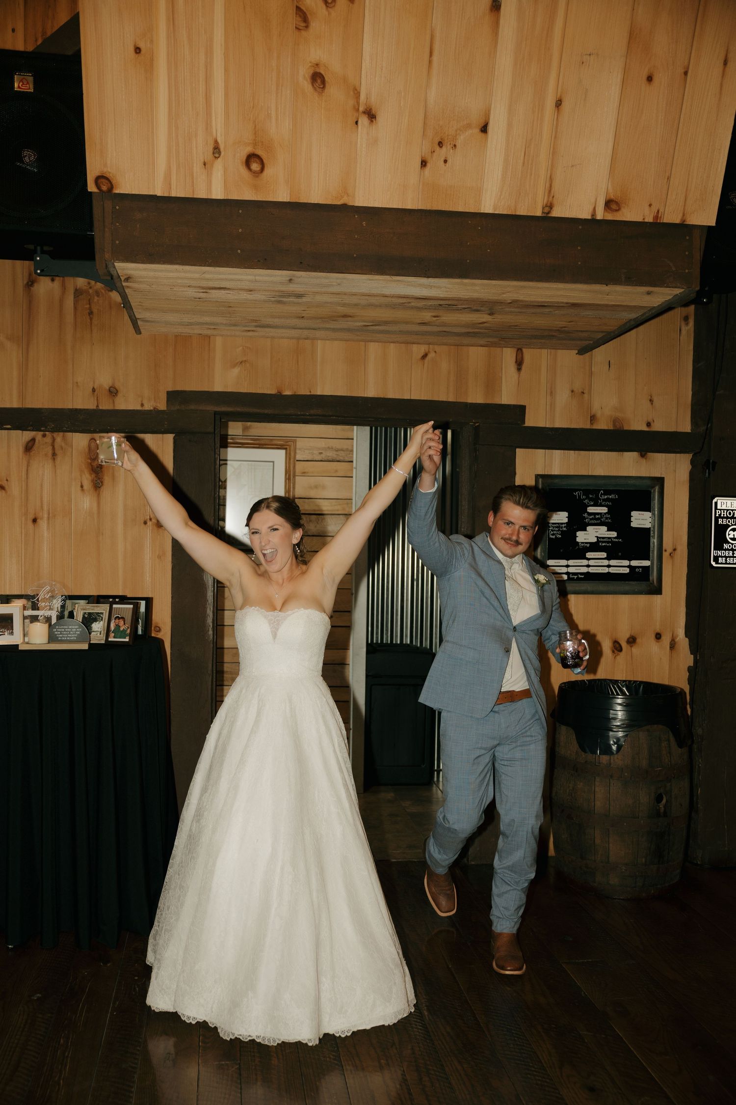 Bride in a white gown and groom in a light blue suit raise their joined hands as they enter a rustic wooden barn wedding reception.