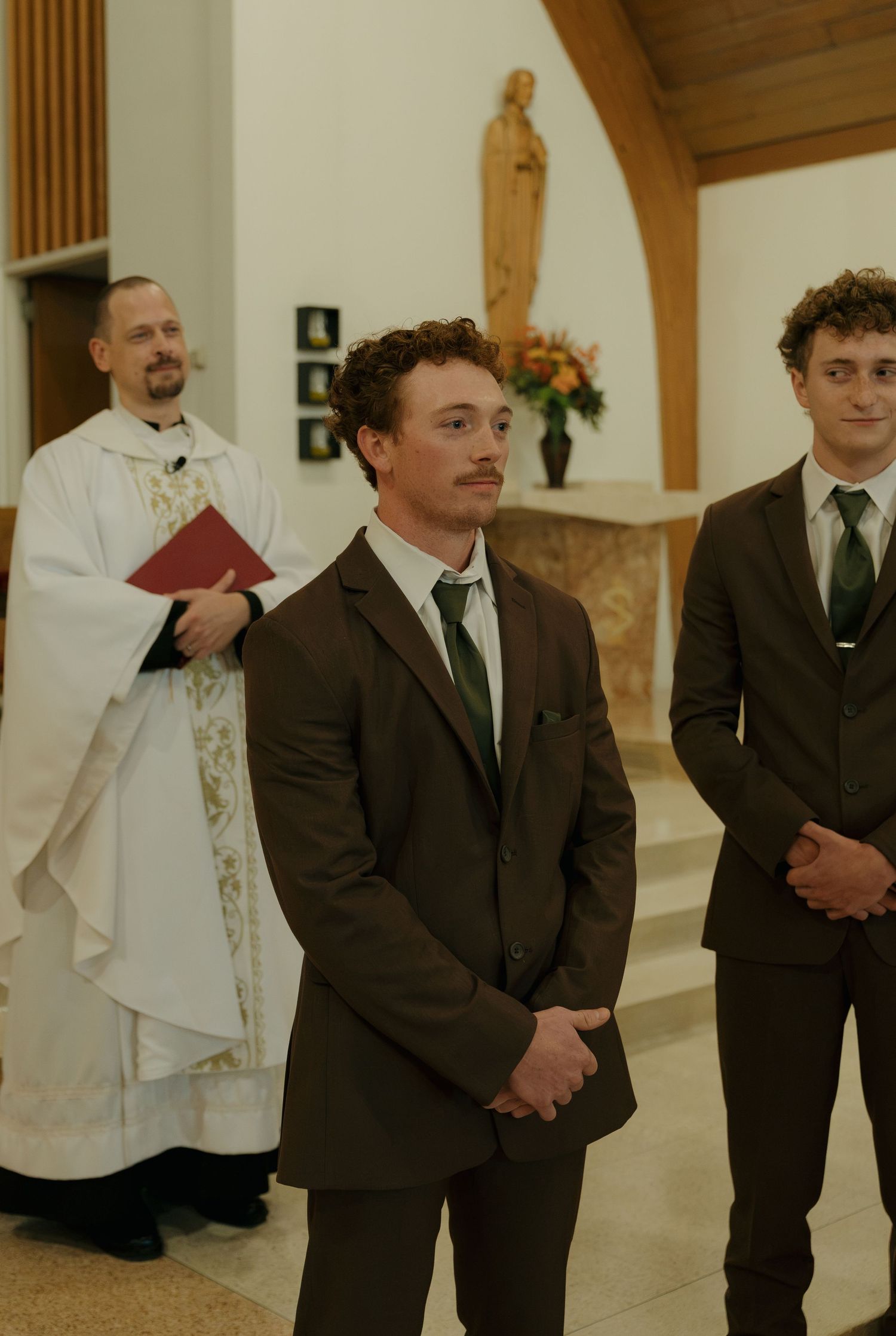 Two groomsmen in brown suits and green ties stand with hands clasped as a priest in white vestments holds a red book in a church.