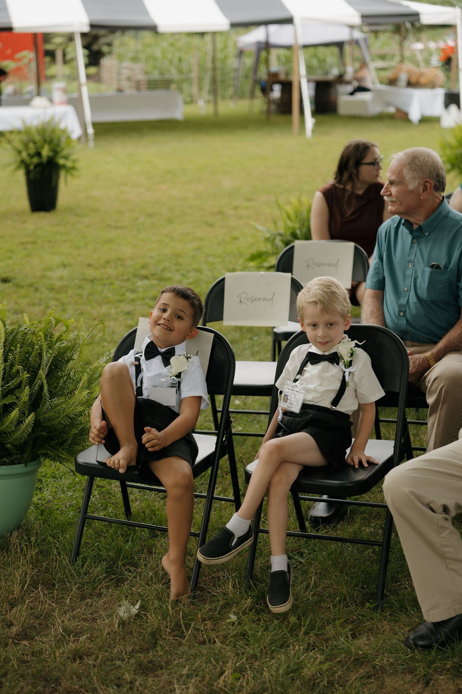 Two young ring bearers in suspenders and bow ties sit on black folding chairs at an outdoor wedding ceremony.