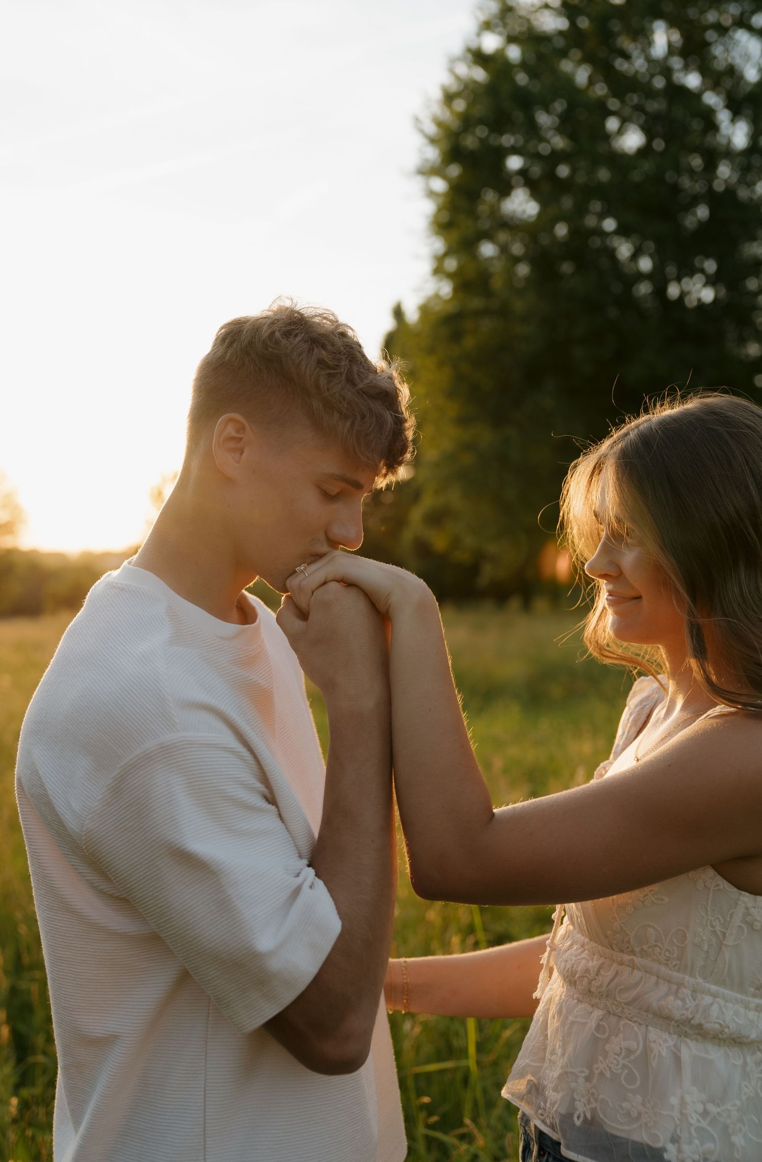A young couple shares a romantic moment at golden hour, with the man kissing the woman's hand in a sunlit field.