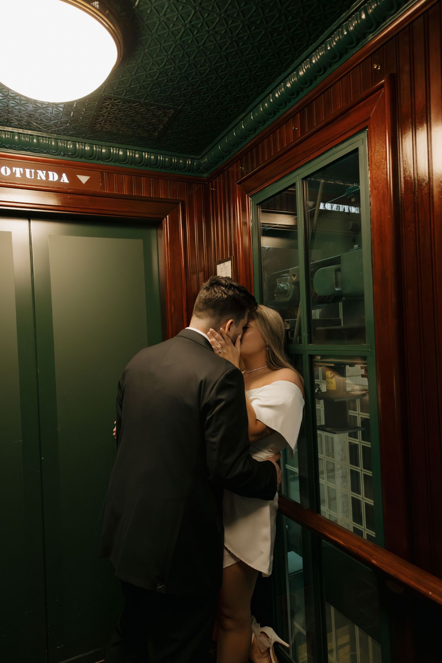 Couple sharing a romantic kiss in an elegant green and wood-paneled elevator, woman in white dress, man in dark suit.