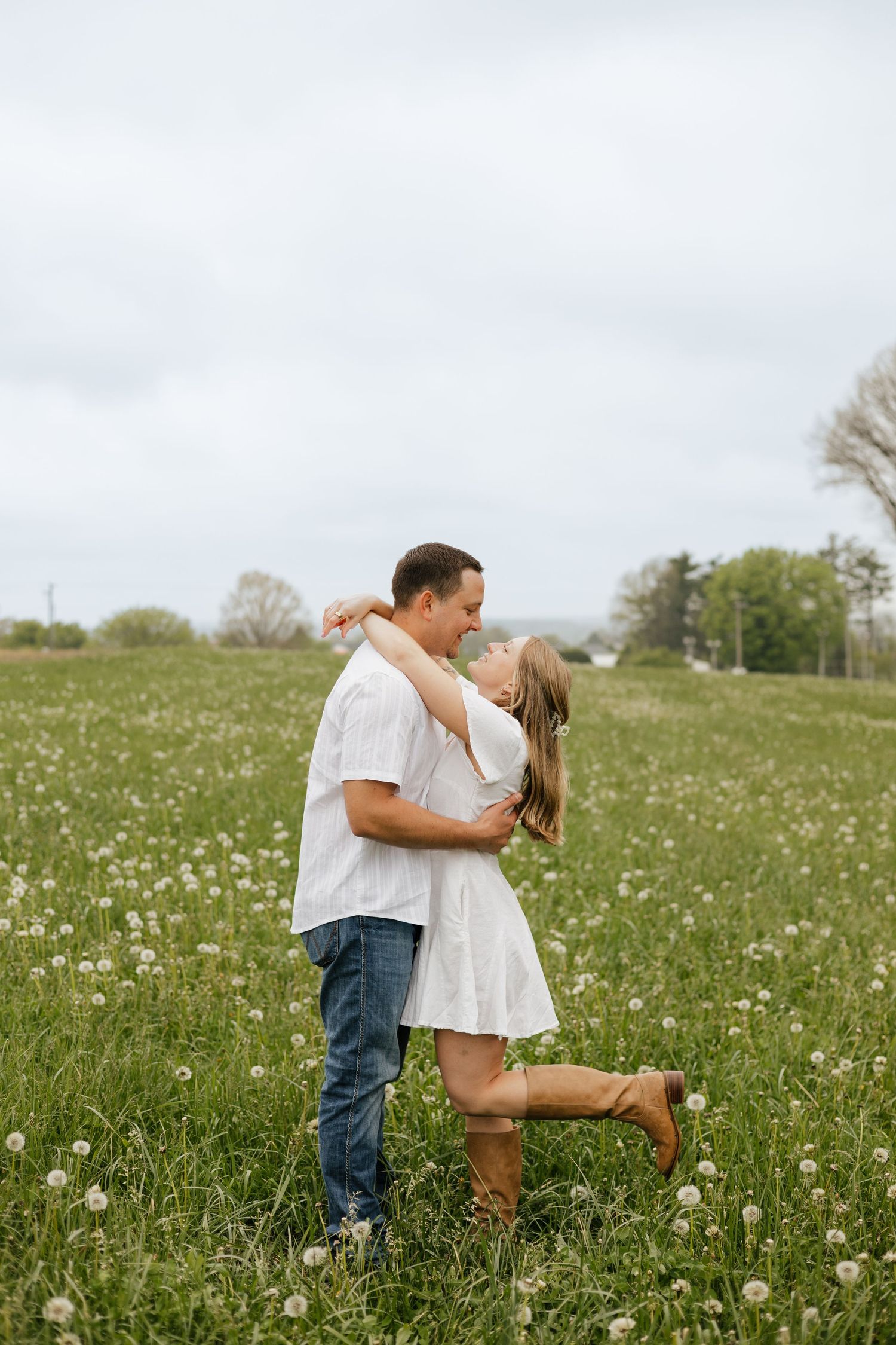 Couple embracing and about to kiss in a lush green field of dandelions, woman in white dress and brown boots.