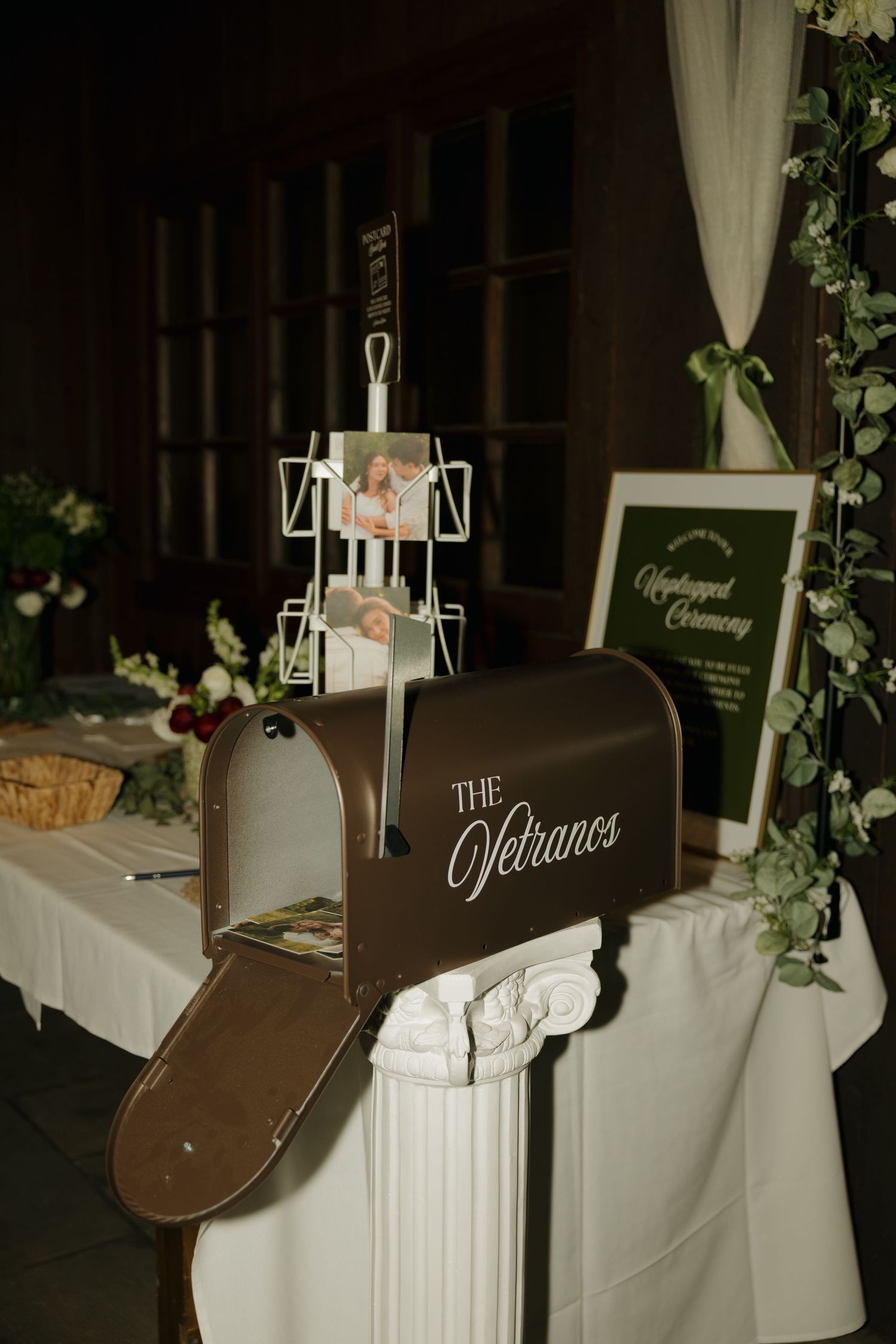 Brown mailbox labeled The Vetranos displayed on white pedestal at elegant wedding card table with greenery decor.