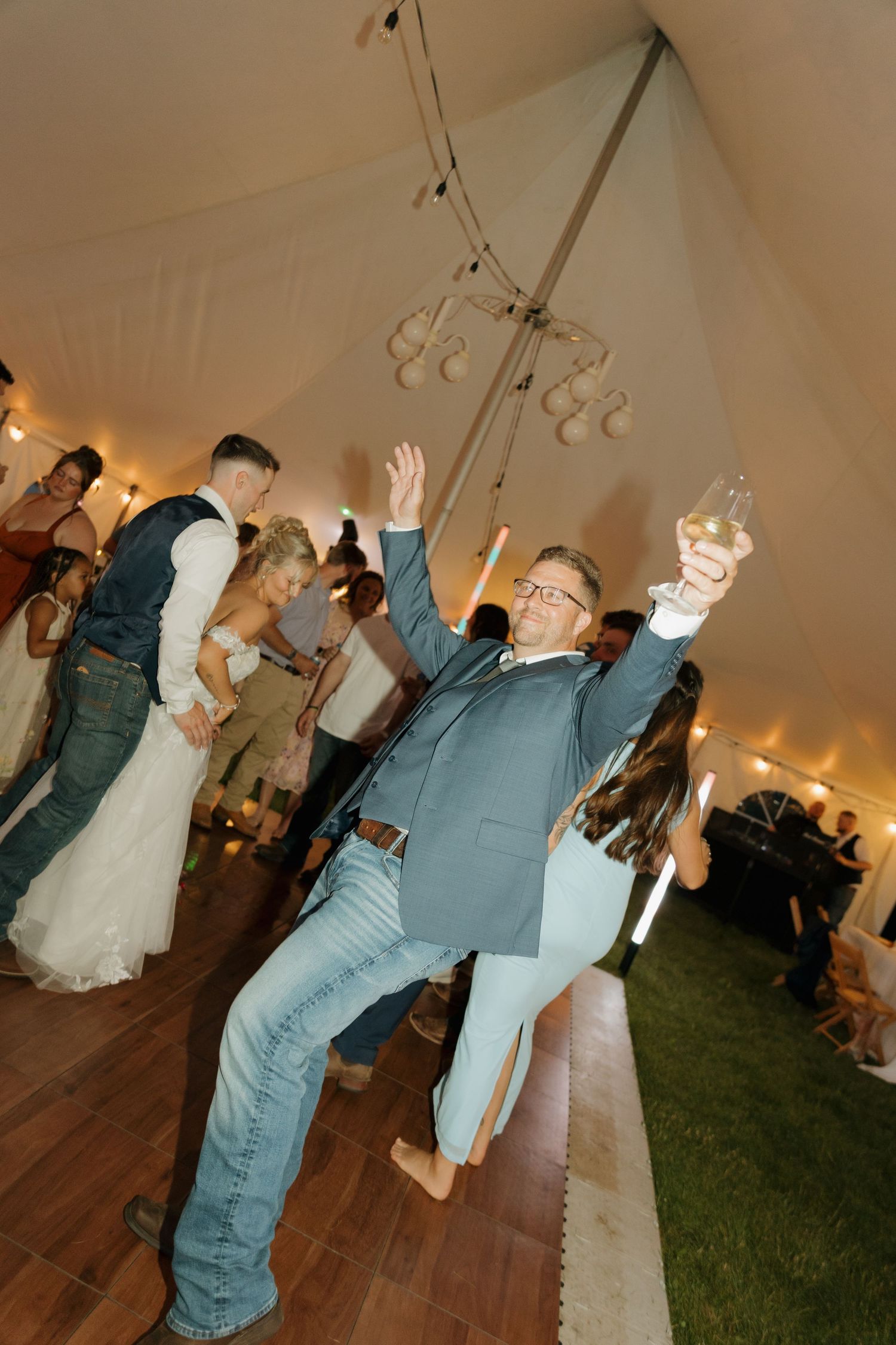 Enthusiastic wedding guest dancing and raising glass under a white reception tent with string lights.