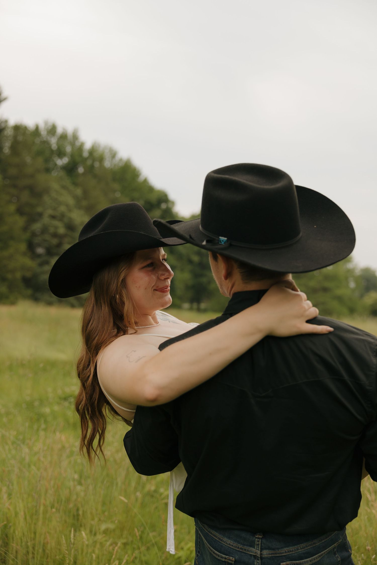 Couple in matching black cowboy hats embrace in a green field, sharing a romantic moment outdoors.