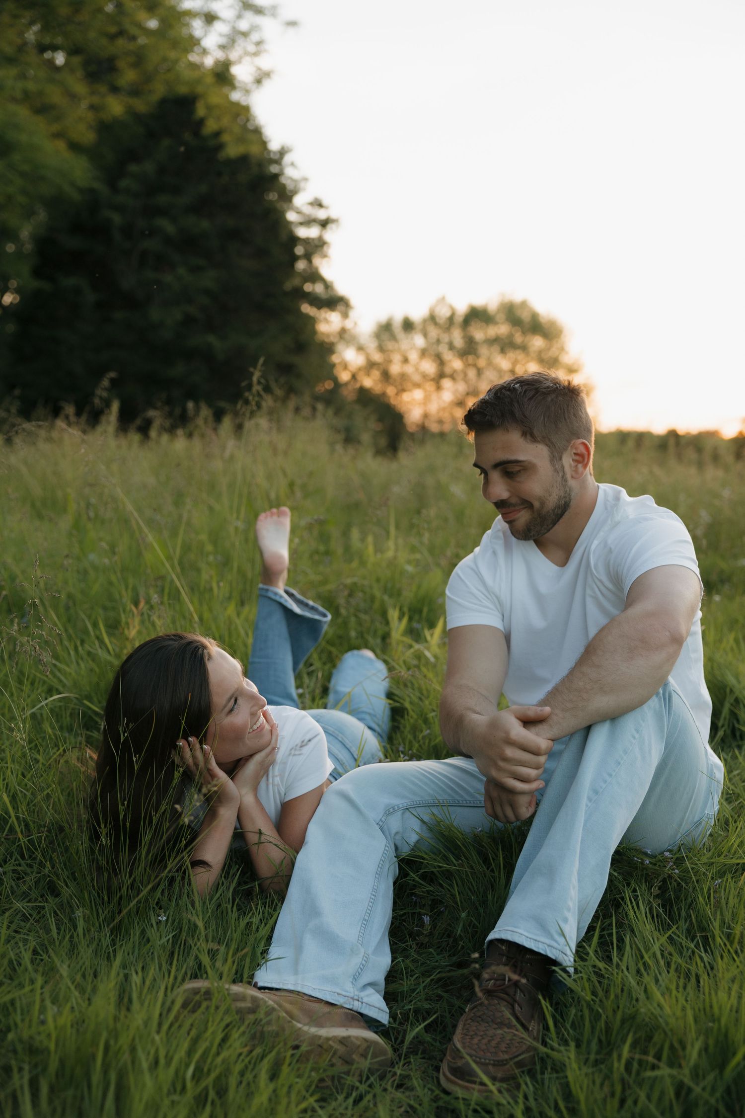 Couple relaxing in green meadow at sunset, woman lying in grass, man sitting beside her, both wearing white outfits.