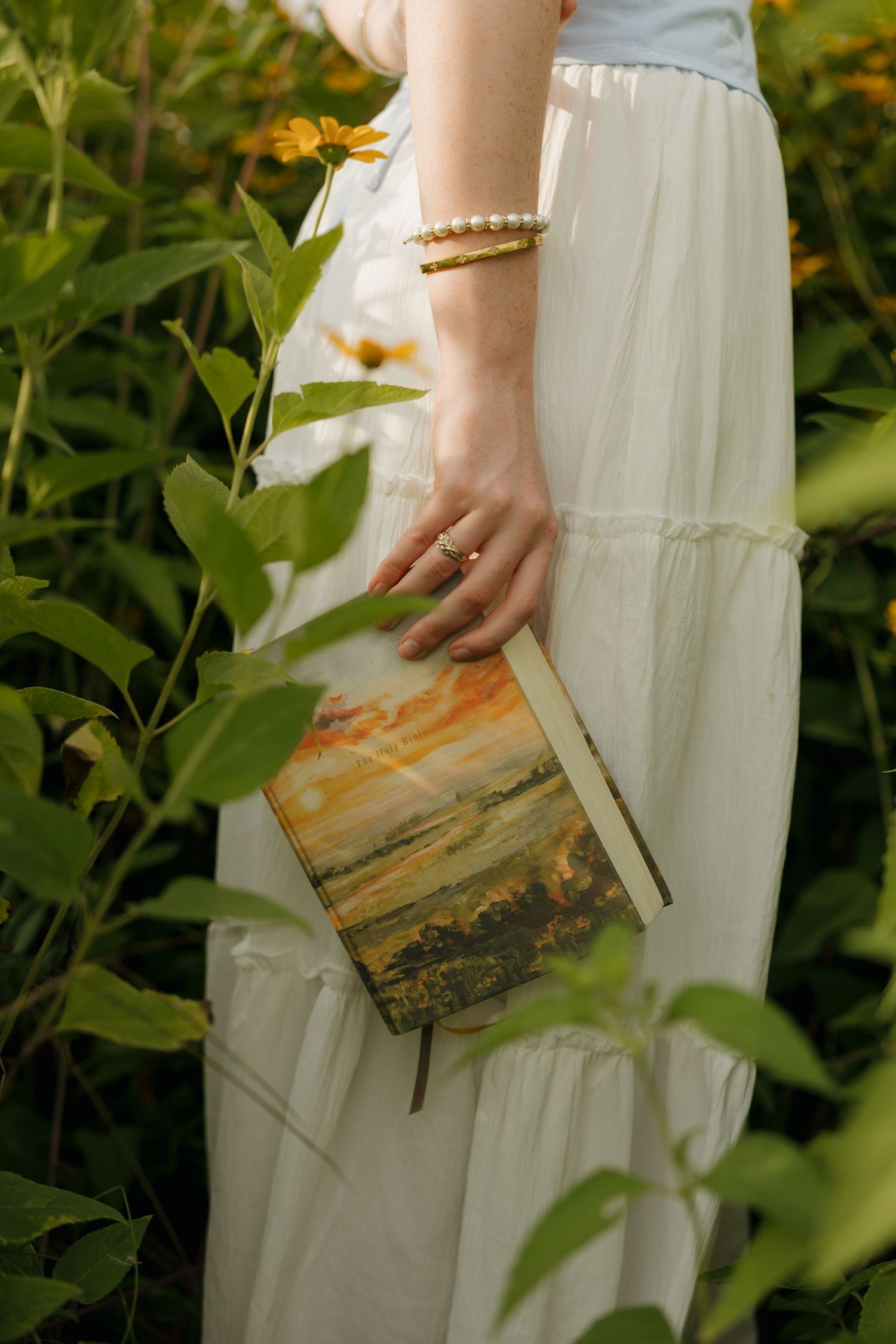 Woman in white skirt holding a landscape-painted journal among sunflowers, wearing gold pearl bracelet and ring.