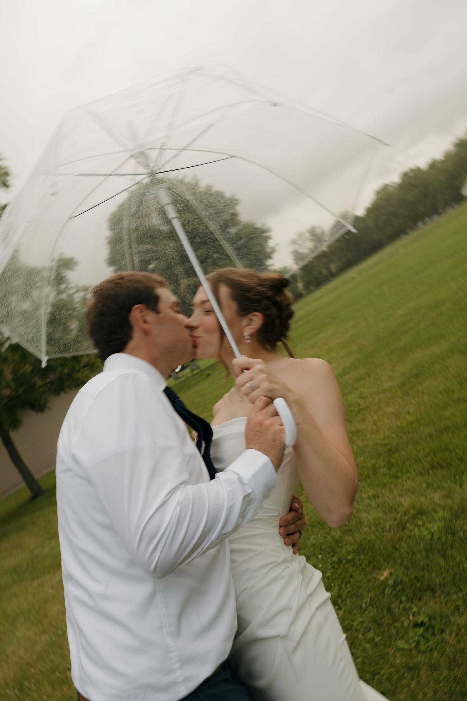 Bride and groom kissing under a clear umbrella on a rainy day in a lush green park during their wedding.
