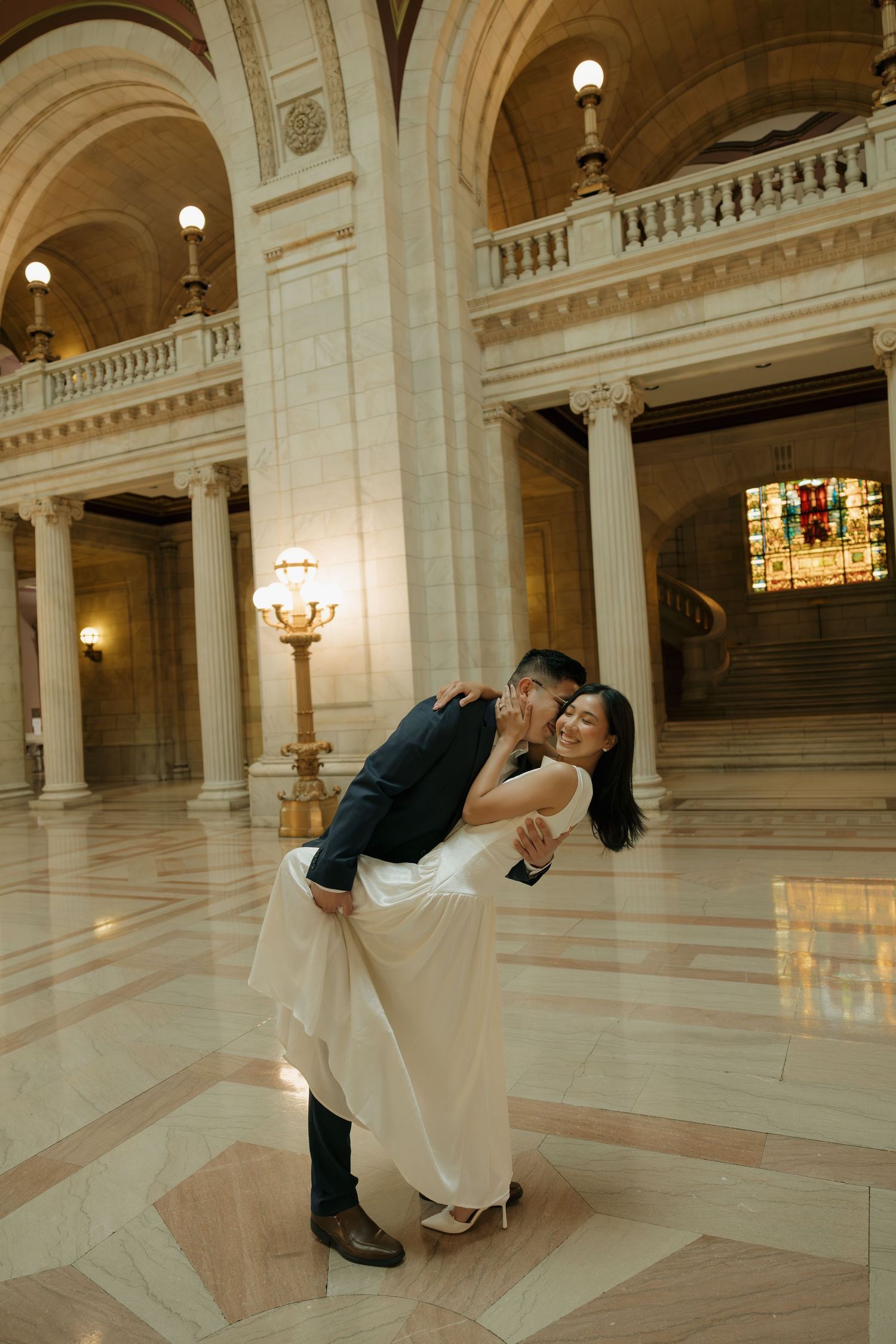 Couple sharing a romantic dip kiss in an elegant marble hall with ornate columns and warm lighting.