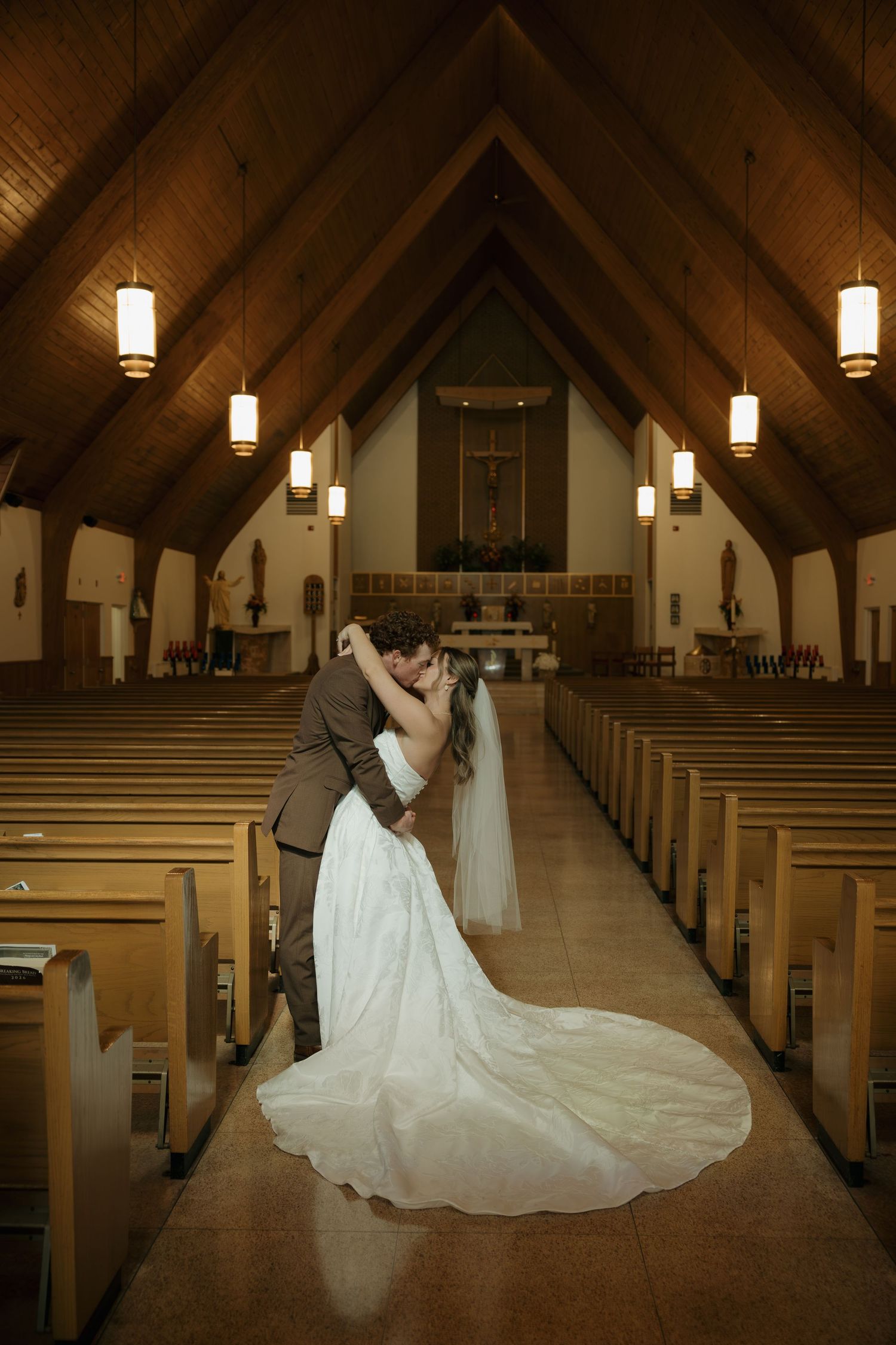 Bride and groom share a dramatic dipped kiss in the aisle of a wooden-beamed church, her long white gown and veil flowing behind her.