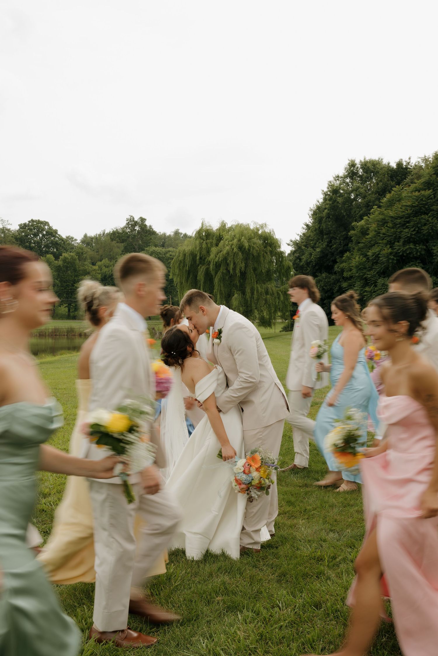 Bride and groom share a kiss surrounded by colorful bridesmaids and groomsmen walking past on a green lawn.