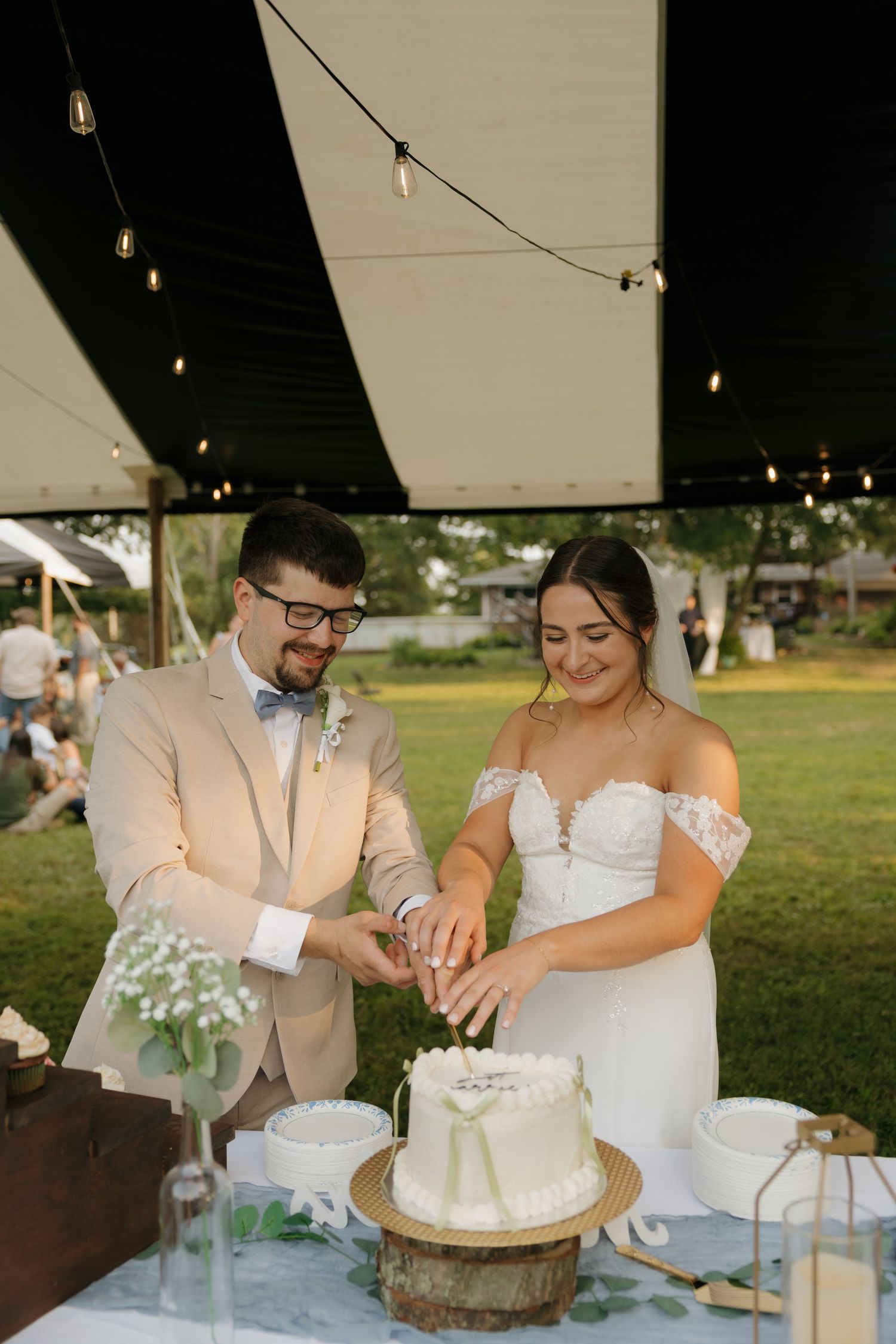 Bride and groom cutting their white wedding cake together under string lights at an outdoor reception.