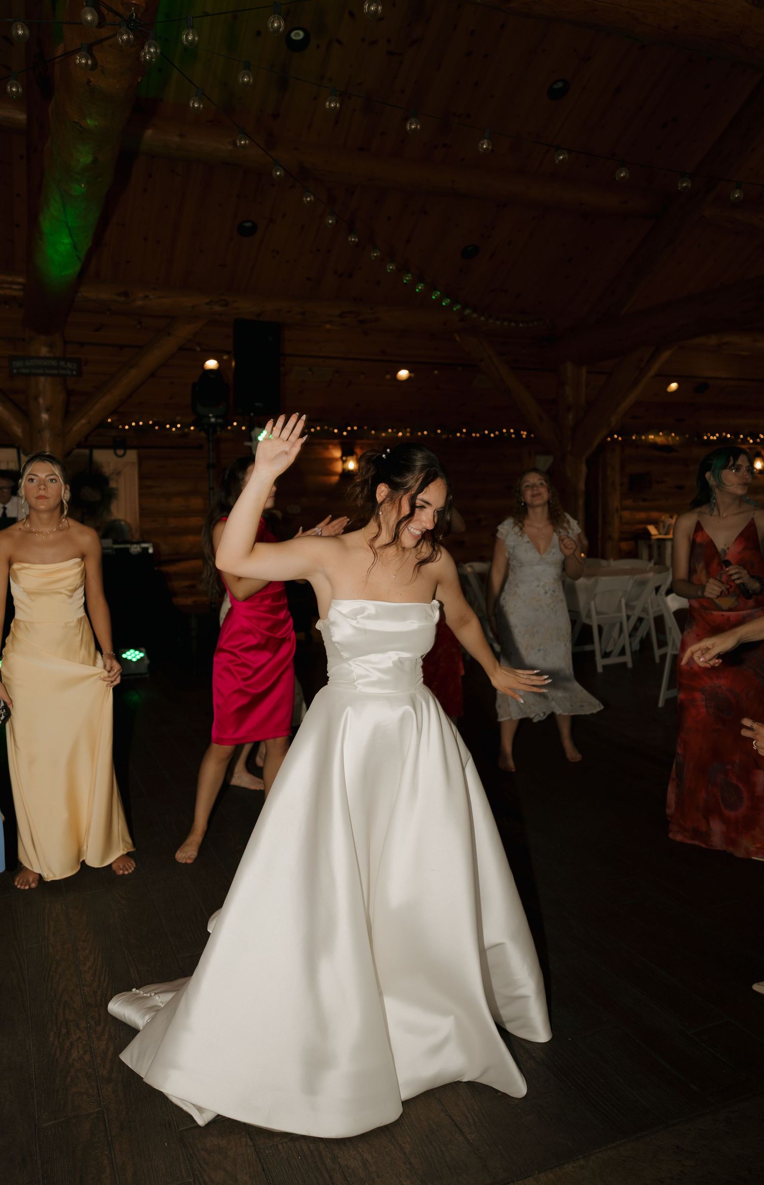 Bride in strapless white satin gown dances joyfully with guests in rustic barn wedding reception venue.