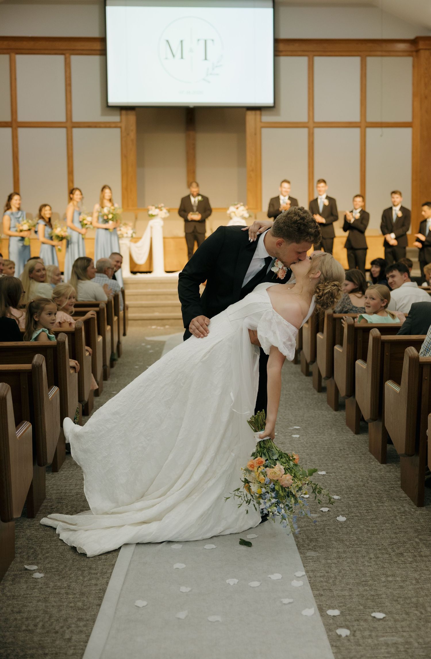 Groom dipping and kissing bride in church aisle after wedding ceremony with guests applauding around them.