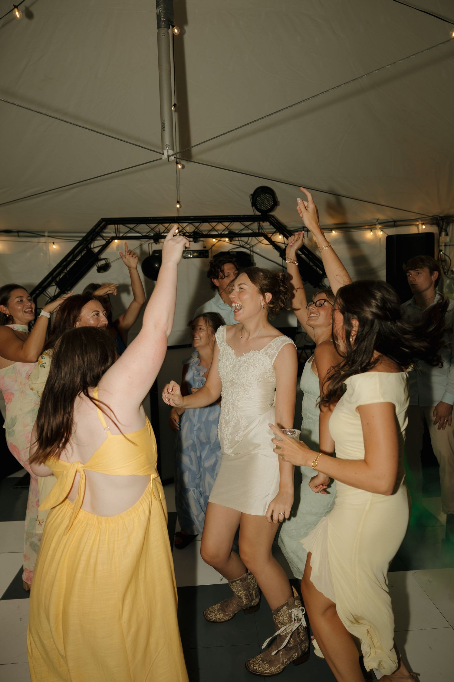 Guests dancing joyfully under a tent at a wedding reception, with the bride in white at the center.
