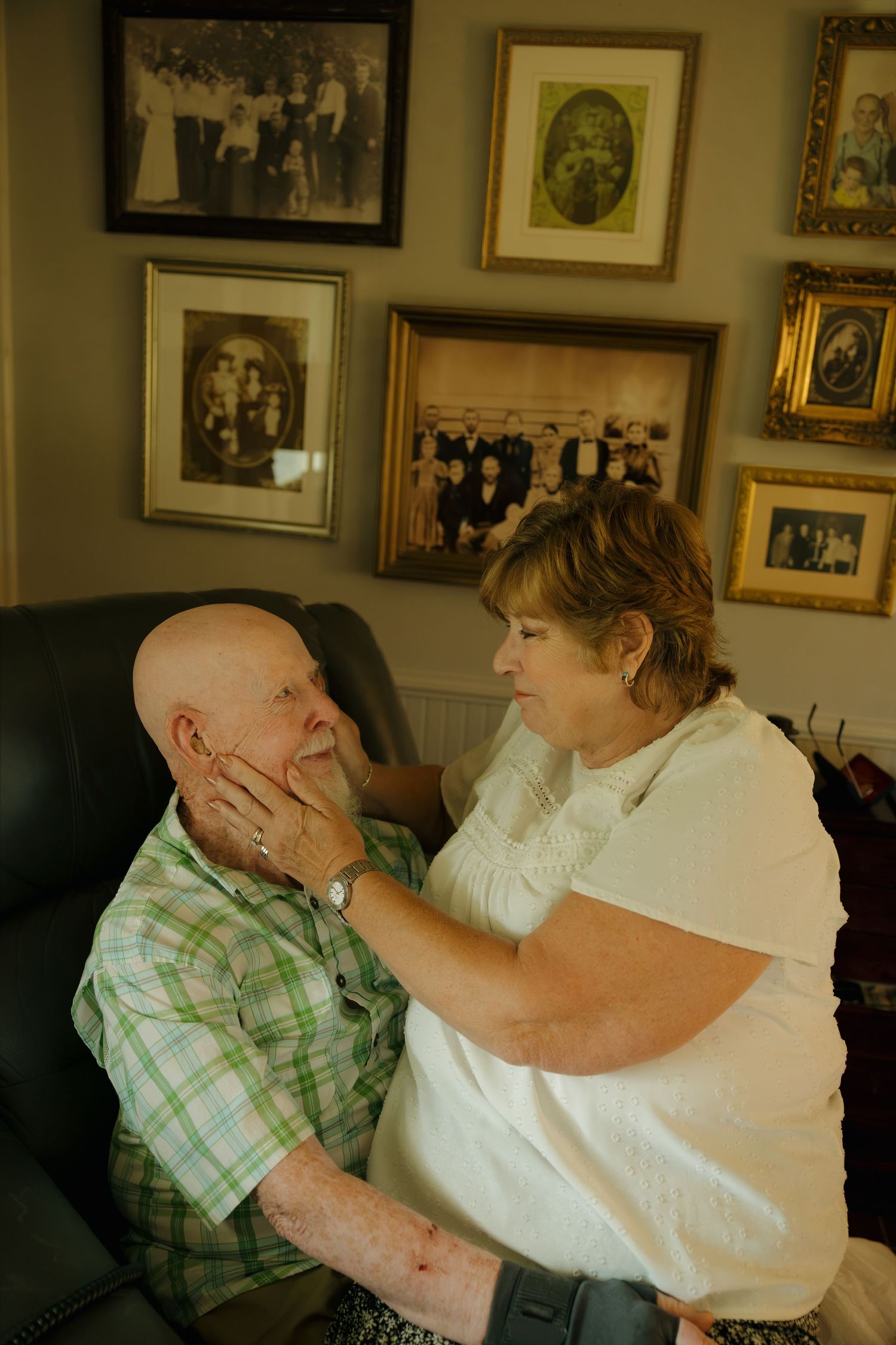 Elderly couple sharing tender moment at home, woman gently holding mans face, family photos displayed on wall behind them.