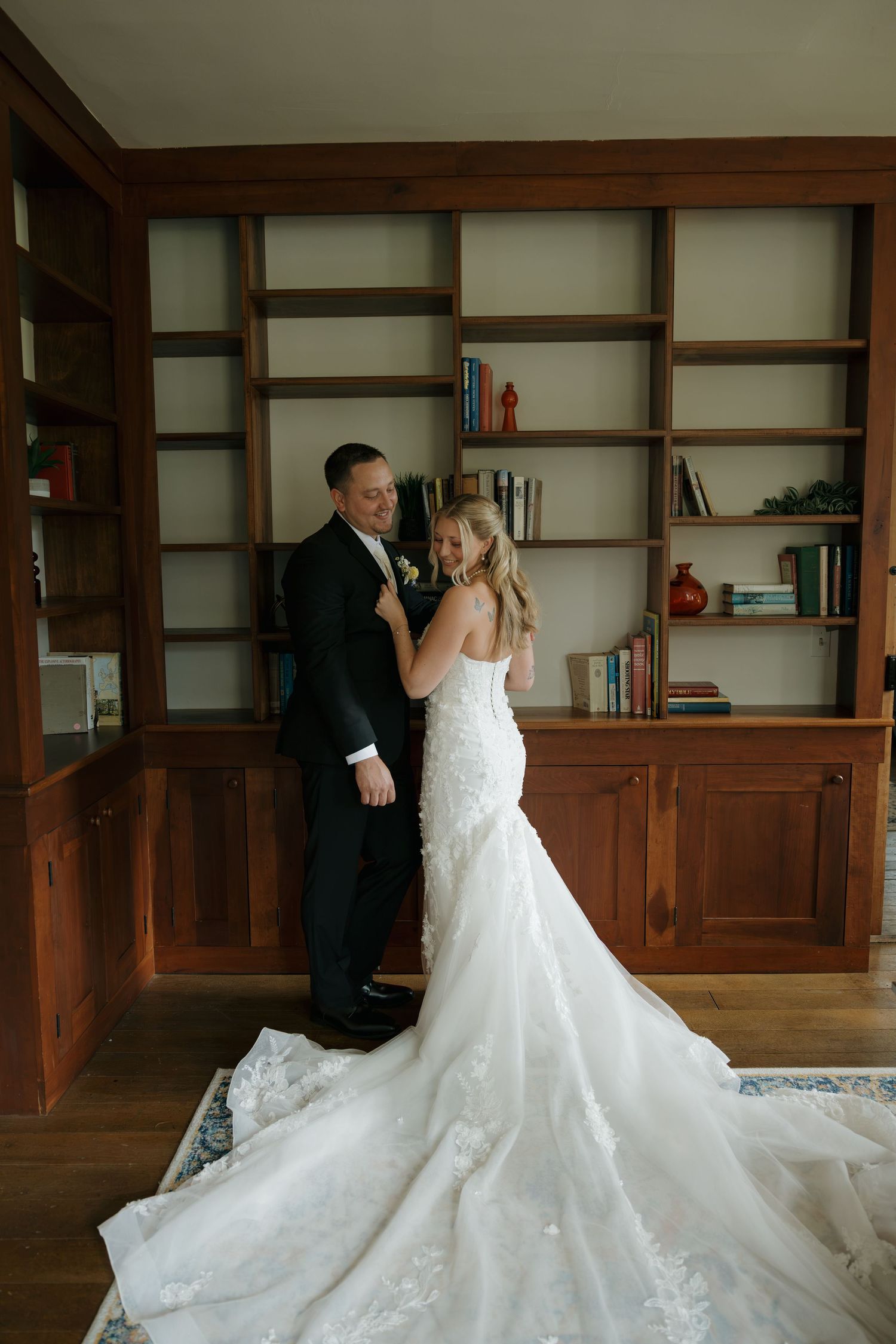 Bride in lace mermaid gown with long train stands with groom in black tuxedo before wooden bookshelves.