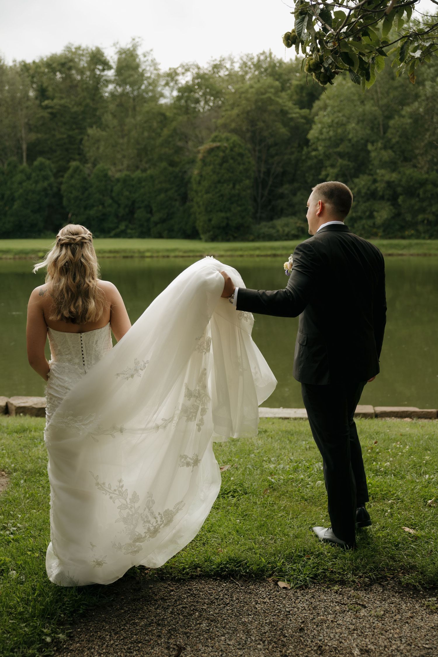 Groom holds bride's flowing lace wedding train as couple walks toward a scenic lake surrounded by trees.