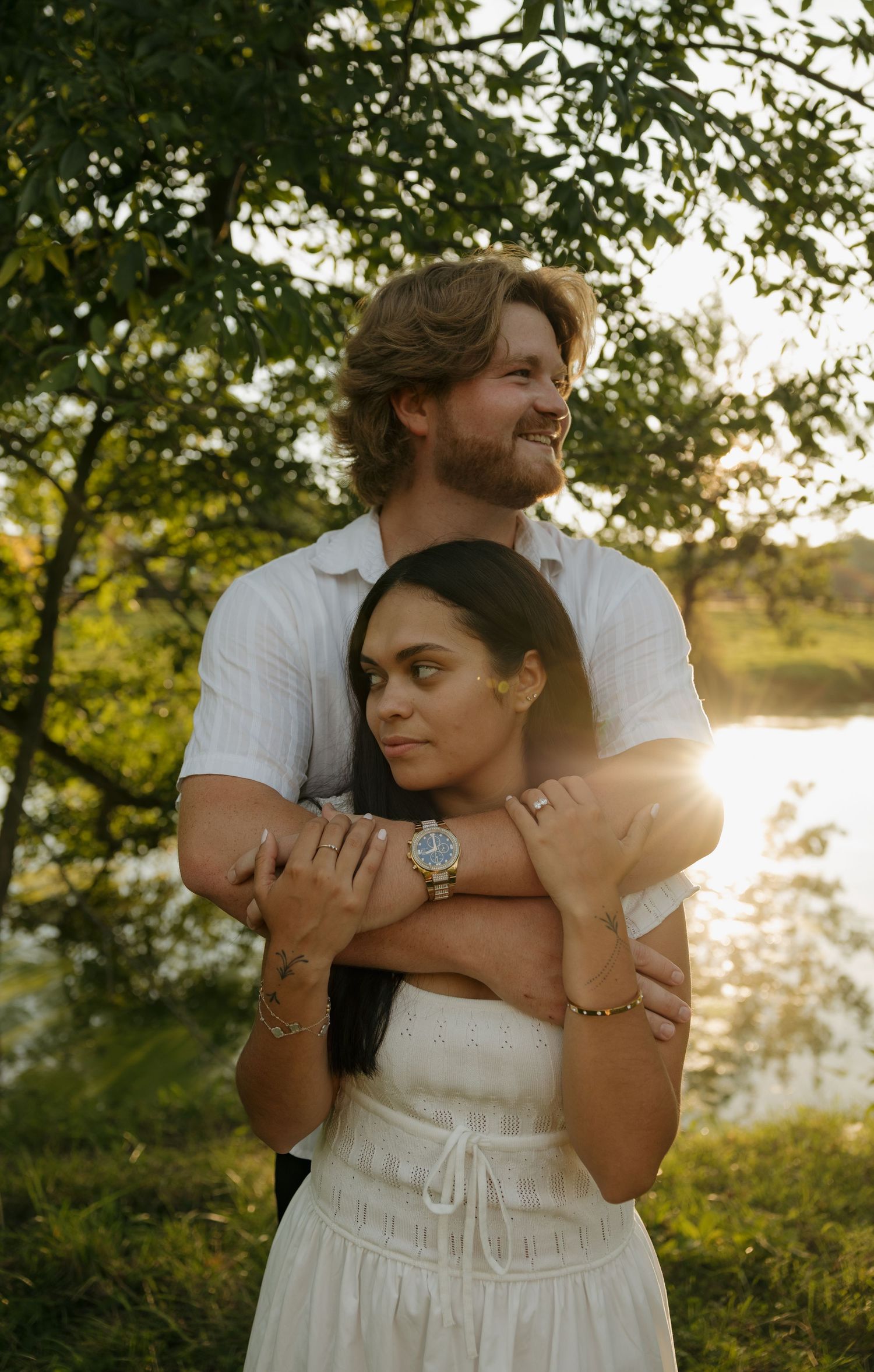 Couple embracing outdoors at golden hour near a lake, surrounded by lush green trees in a romantic portrait.