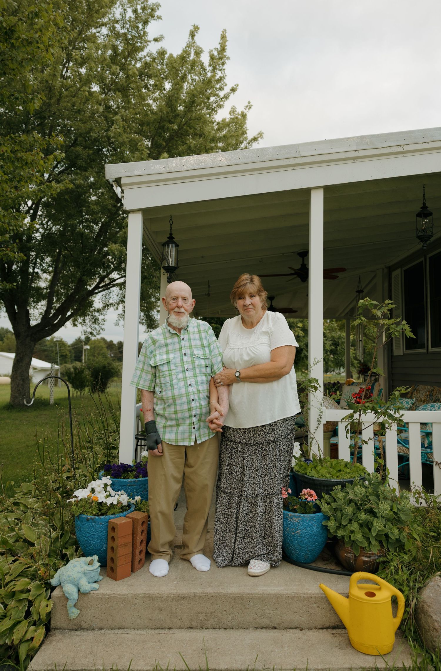 Elderly couple posing together on home porch steps surrounded by colorful potted flowers and garden decorations.
