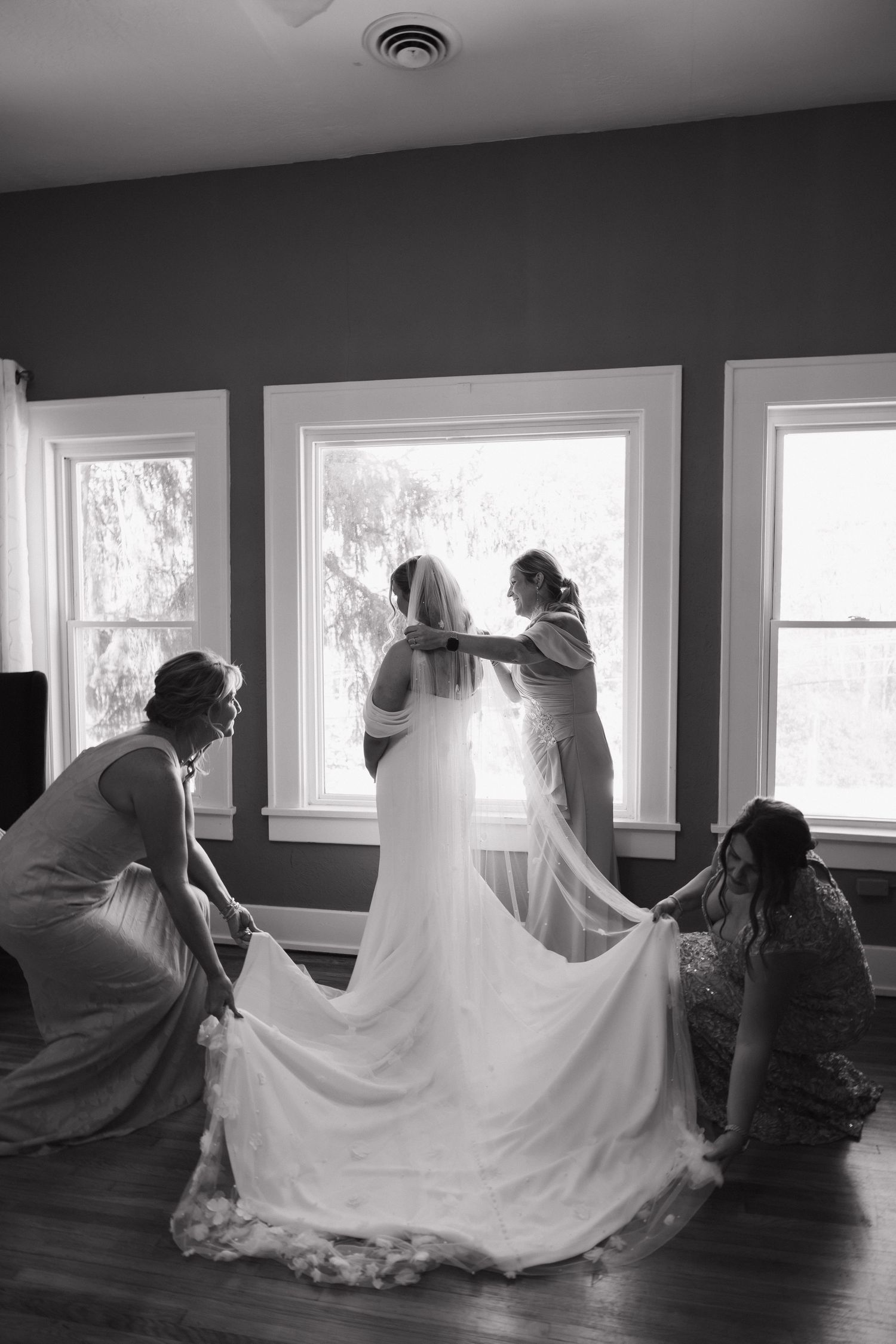 Black and white photo of bridesmaids helping bride with veil and train by sunlit windows before wedding ceremony.