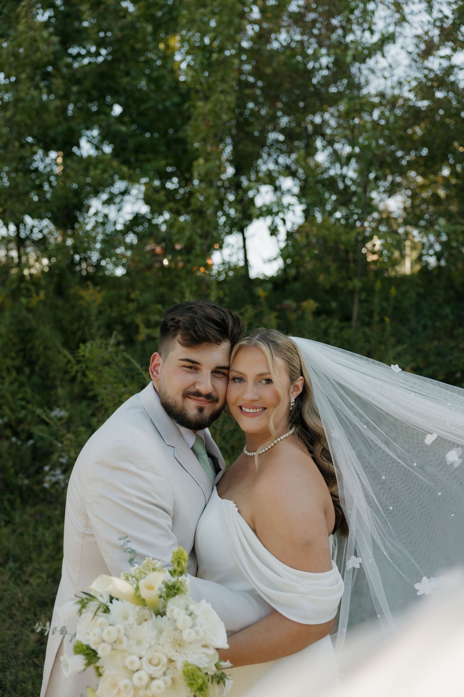 A bride and groom pose outdoors in front of lush green trees, she holds white flowers and wears a flowing veil.