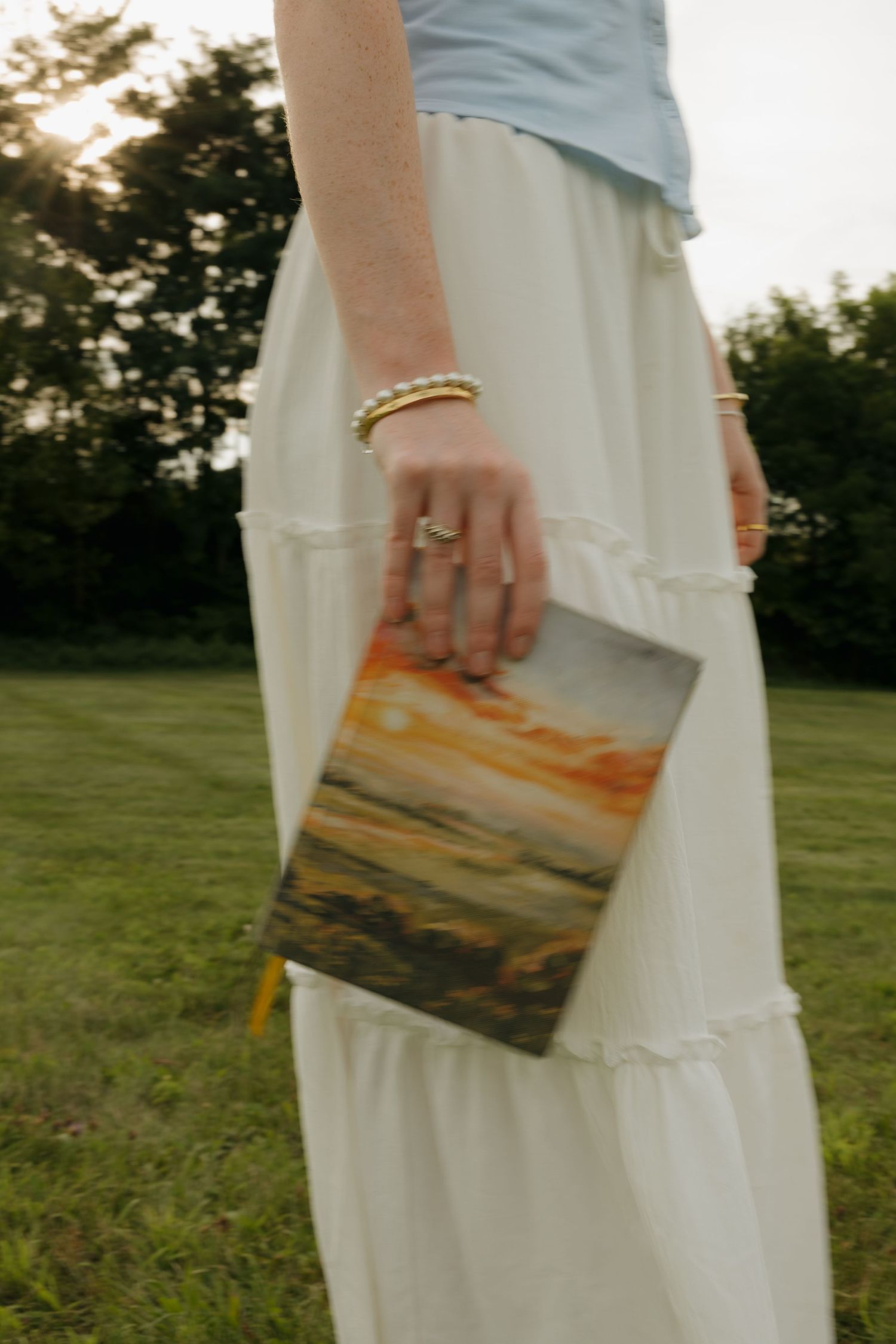 Woman in white maxi skirt holding a colorful landscape painting outdoors on green grass.