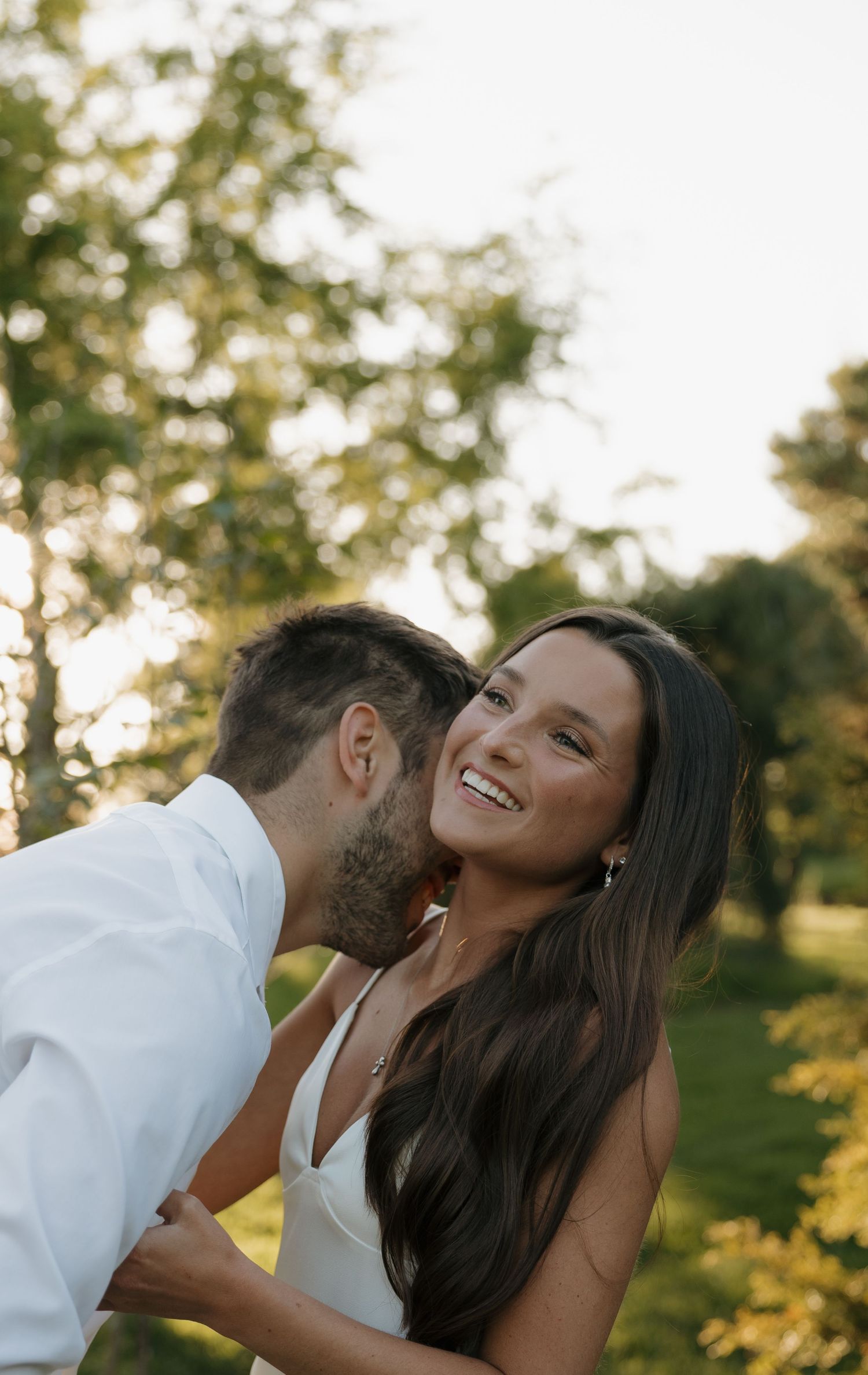 A smiling bride in a white dress receives a kiss on the cheek from a bearded groom outdoors in a sunlit garden.