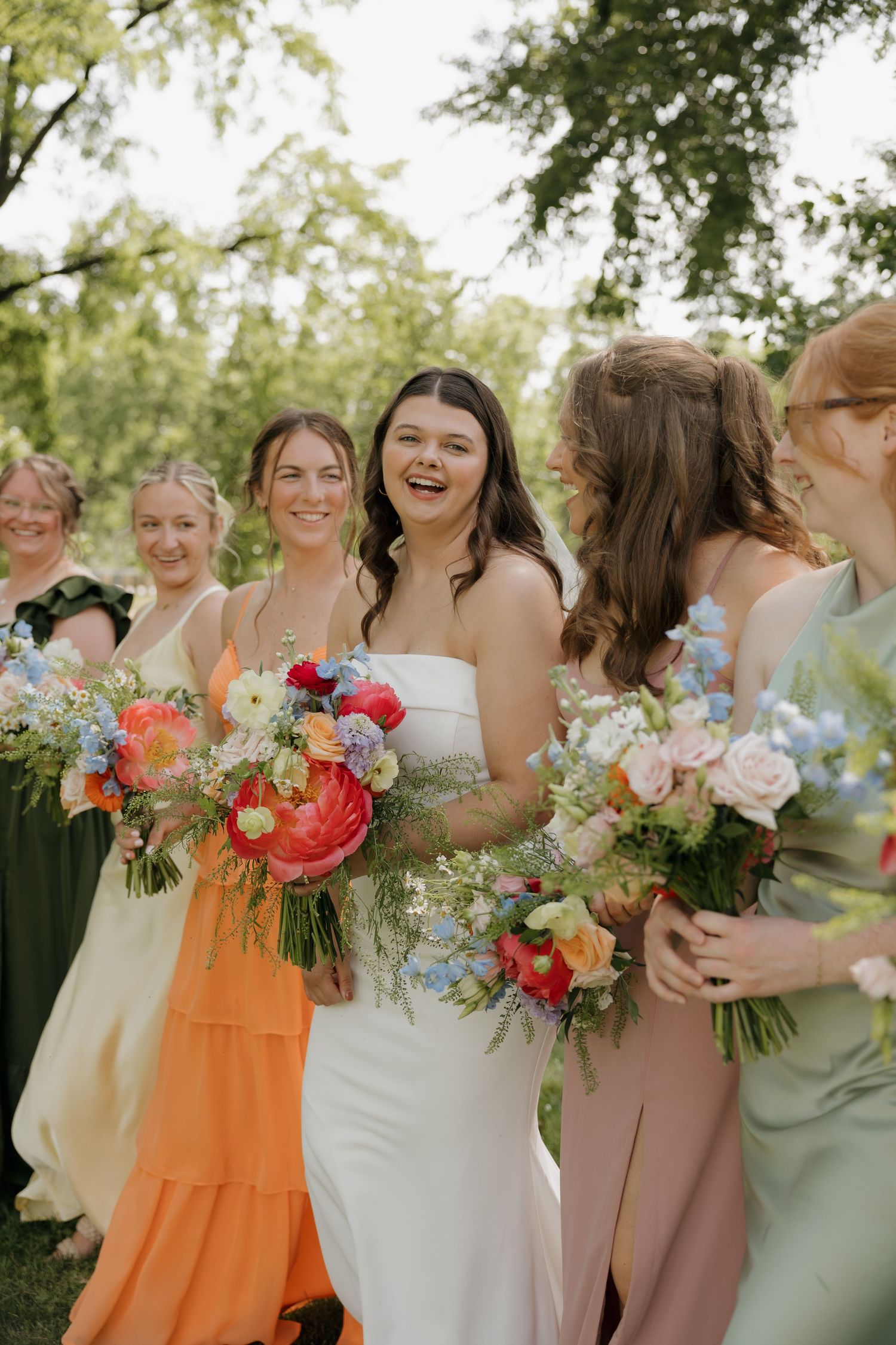 Bride in white strapless gown laughs with bridesmaids in colorful mismatched dresses holding vibrant floral bouquets outdoors.