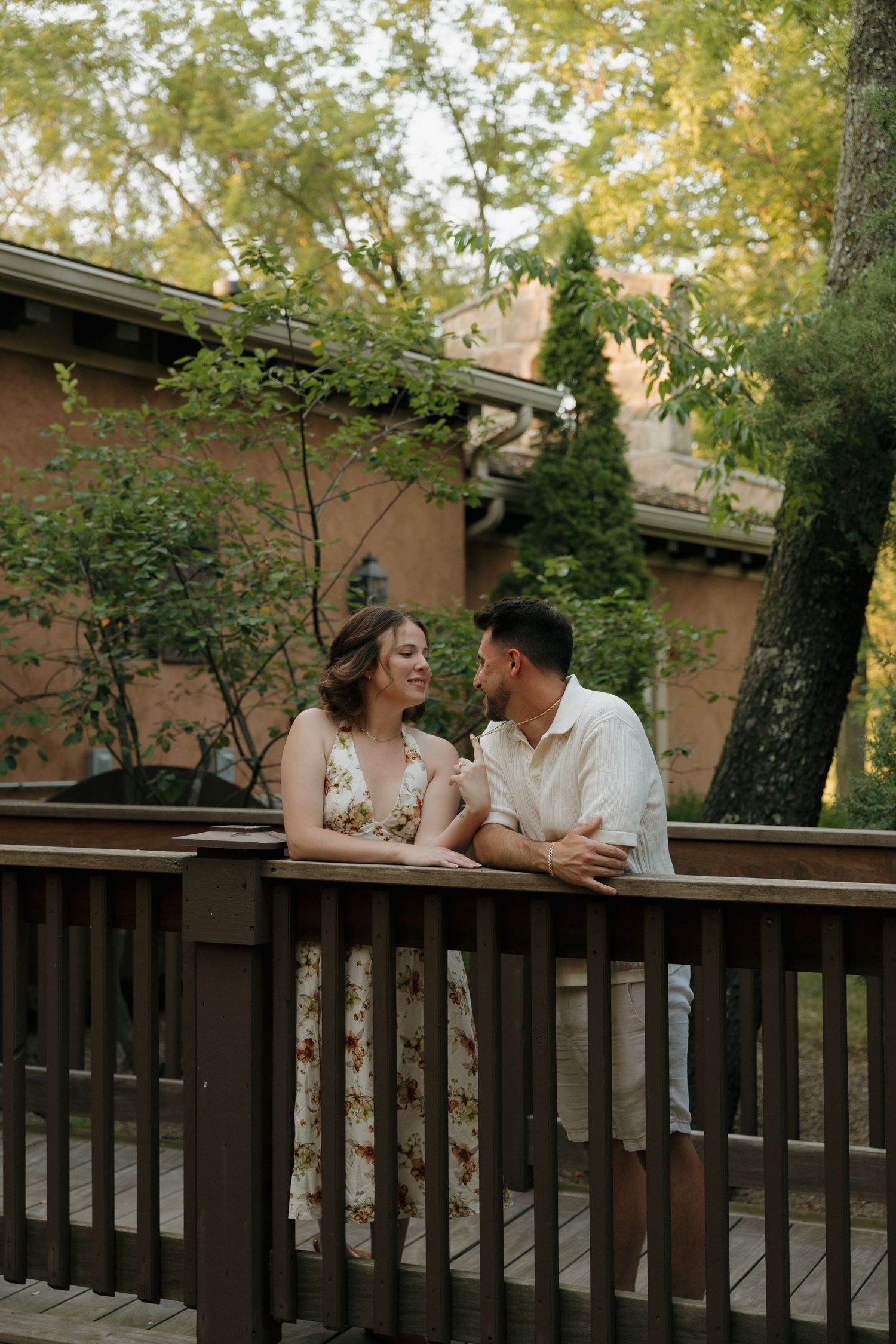 A smiling couple shares a romantic moment on a wooden deck surrounded by lush greenery at golden hour.