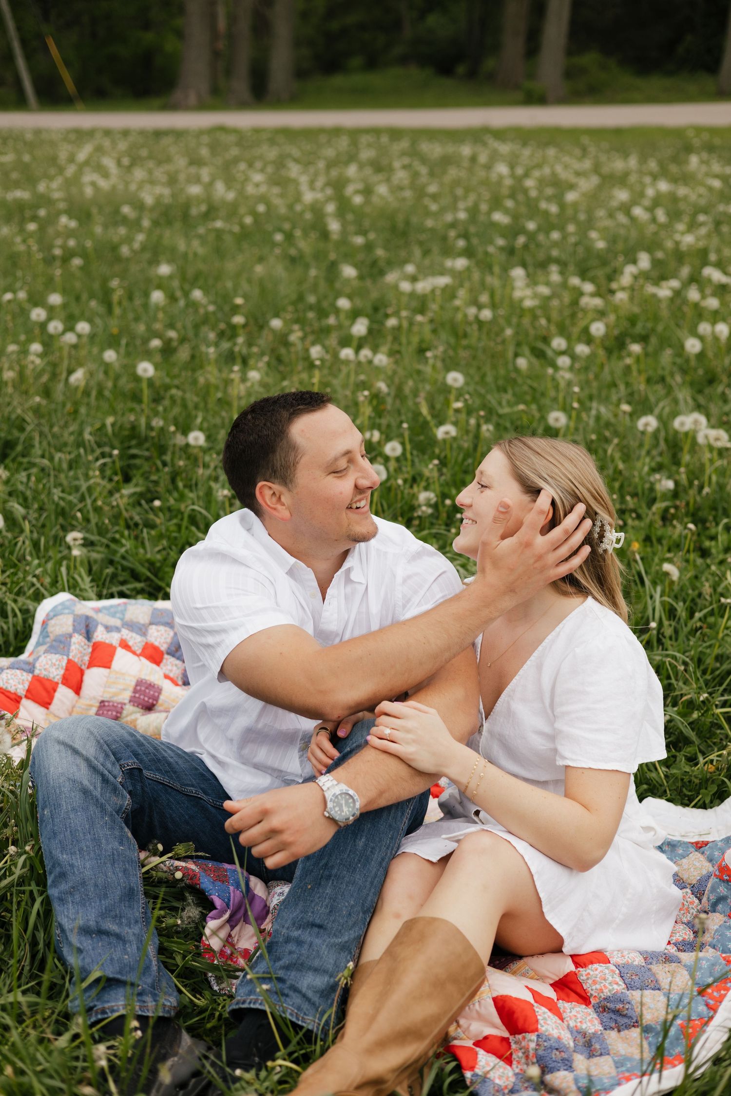 Couple sitting on a colorful quilt in a dandelion field, smiling at each other during a romantic outdoor engagement session.