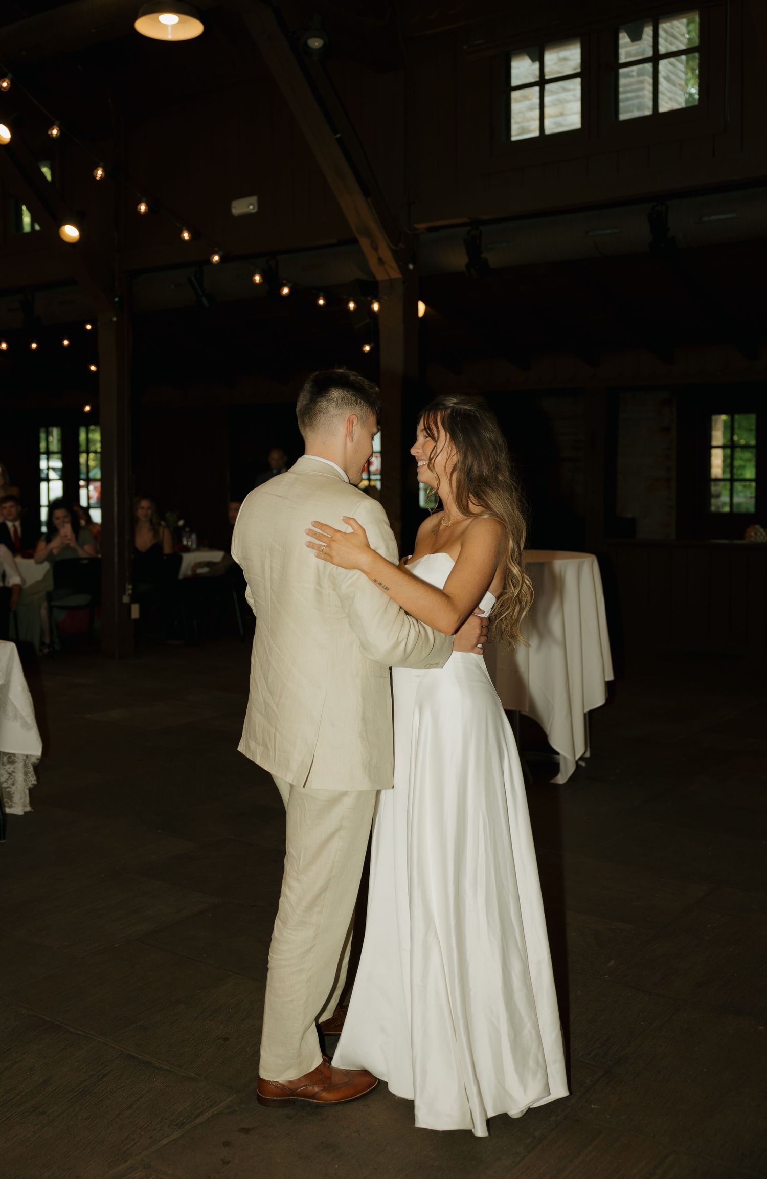 Bride and groom share their first dance in a warmly lit rustic barn venue decorated with string lights.