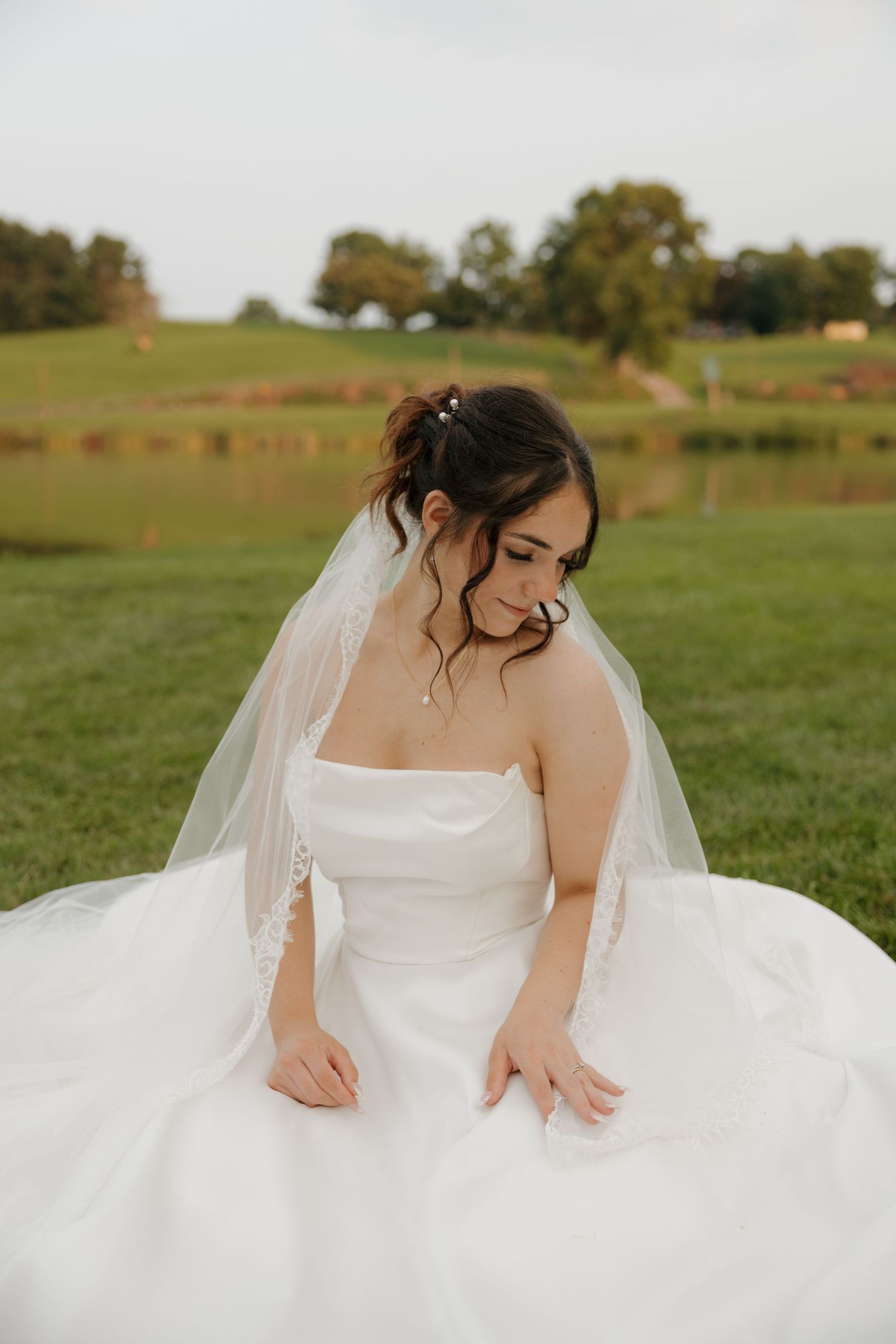 Bride in strapless white gown and veil sits on green grass near a pond, looking down elegantly.