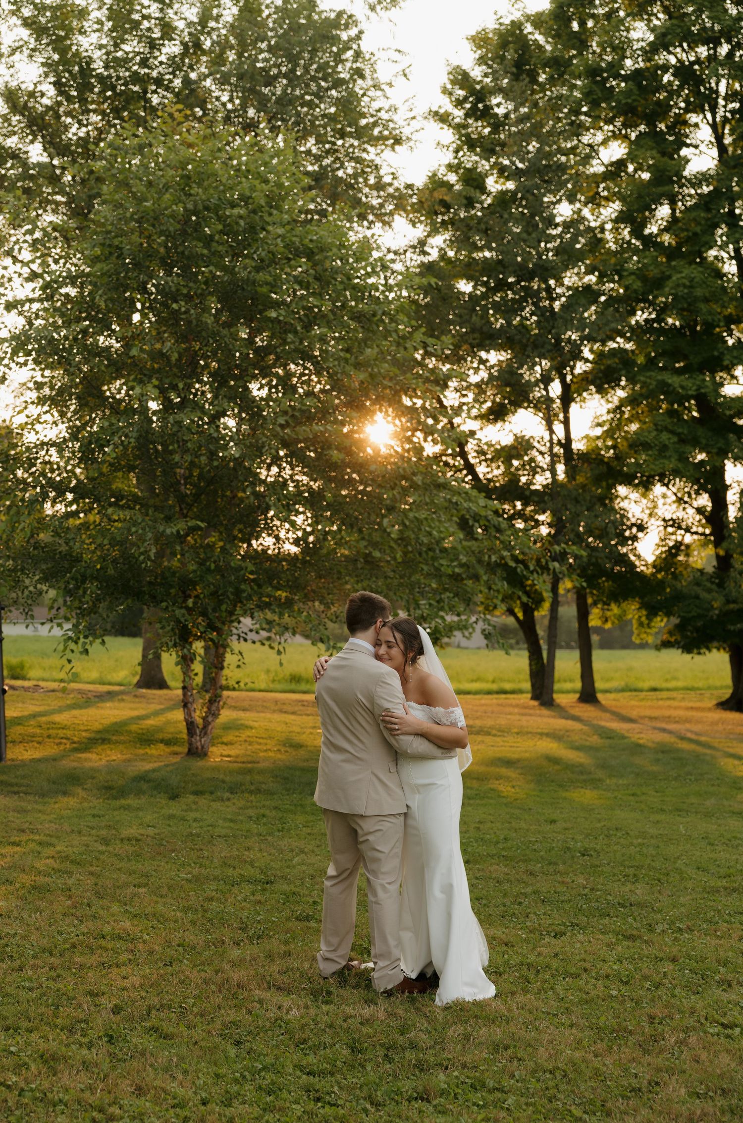 Bride and groom embrace at golden hour sunset in a lush green park during their outdoor wedding celebration.