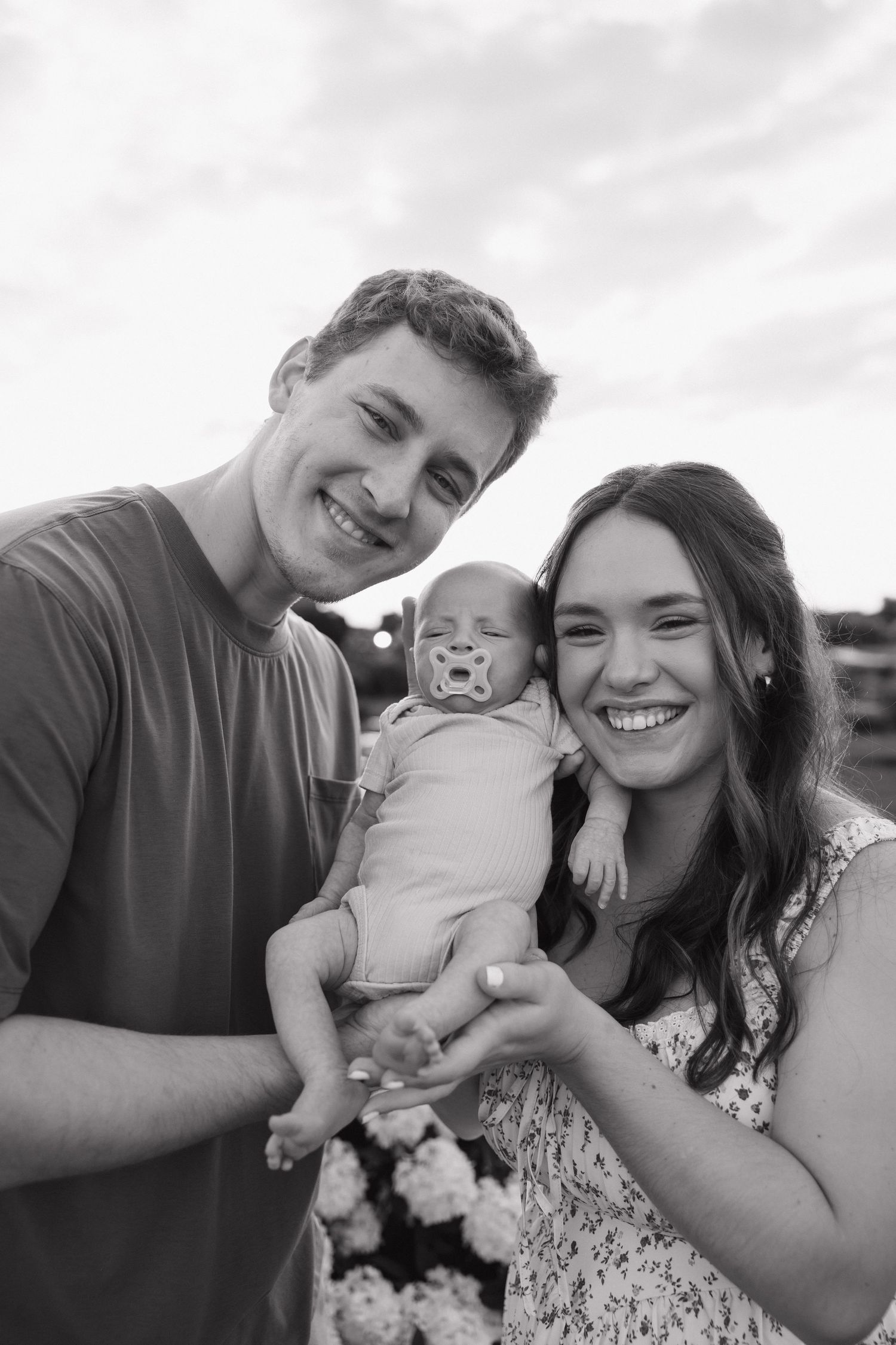 Smiling young parents holding their newborn baby with pacifier outdoors in black and white photo.