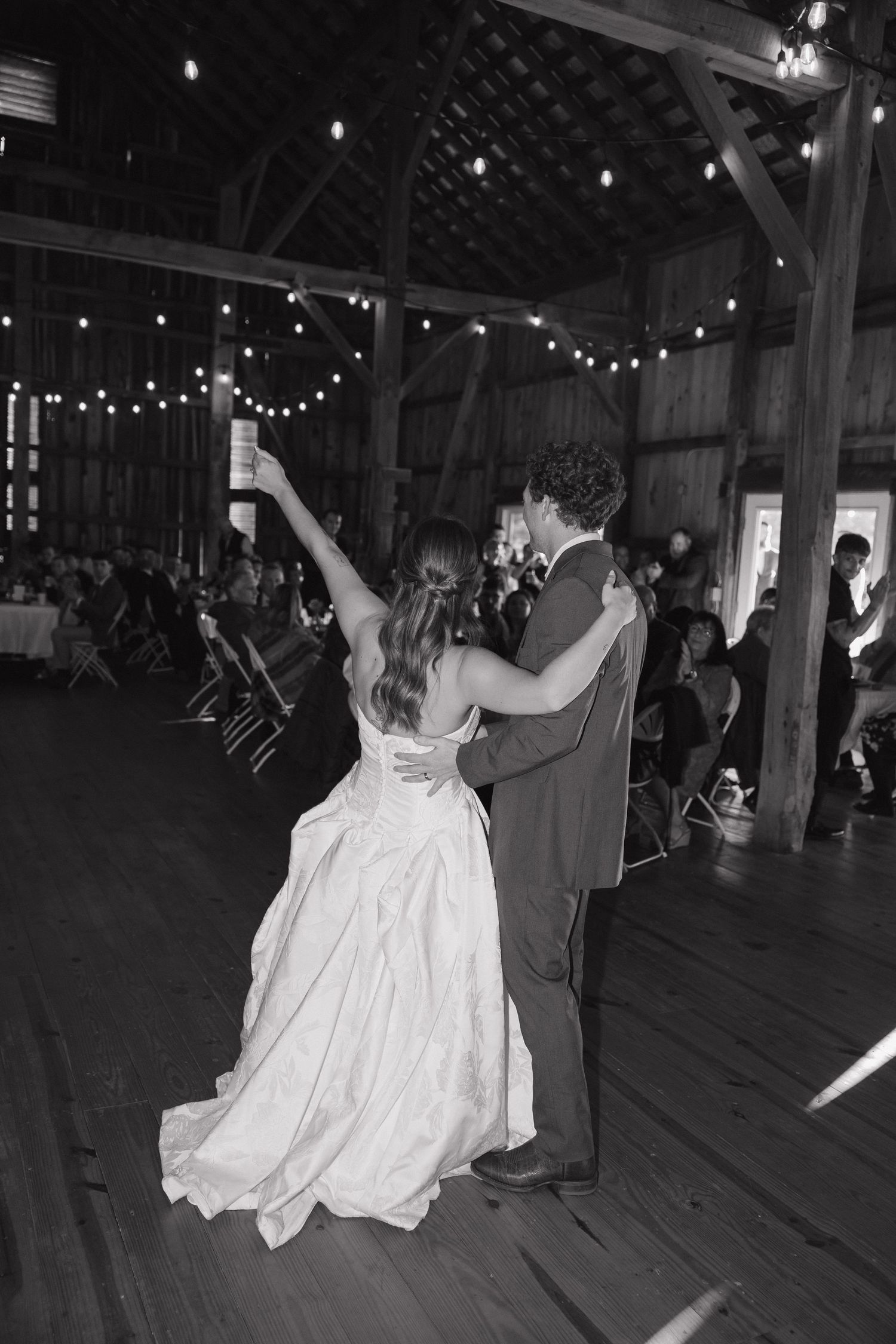 Bride in a flowing gown and groom dance with an arm raised in a string-lit rustic barn as wedding guests look on.