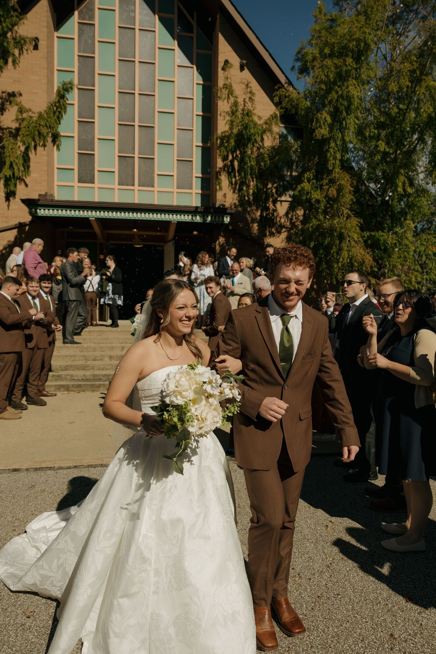 Smiling bride in a strapless white gown holding a bouquet walks arm in arm with the groom in a brown suit past cheering wedding guests.
