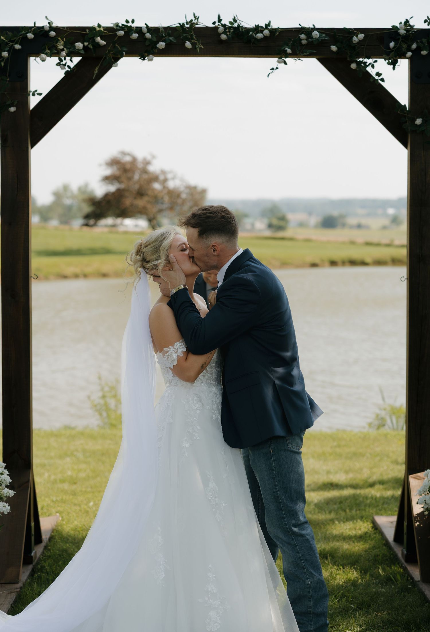 Bride and groom share first kiss under rustic wooden arch decorated with greenery, with scenic pond backdrop.